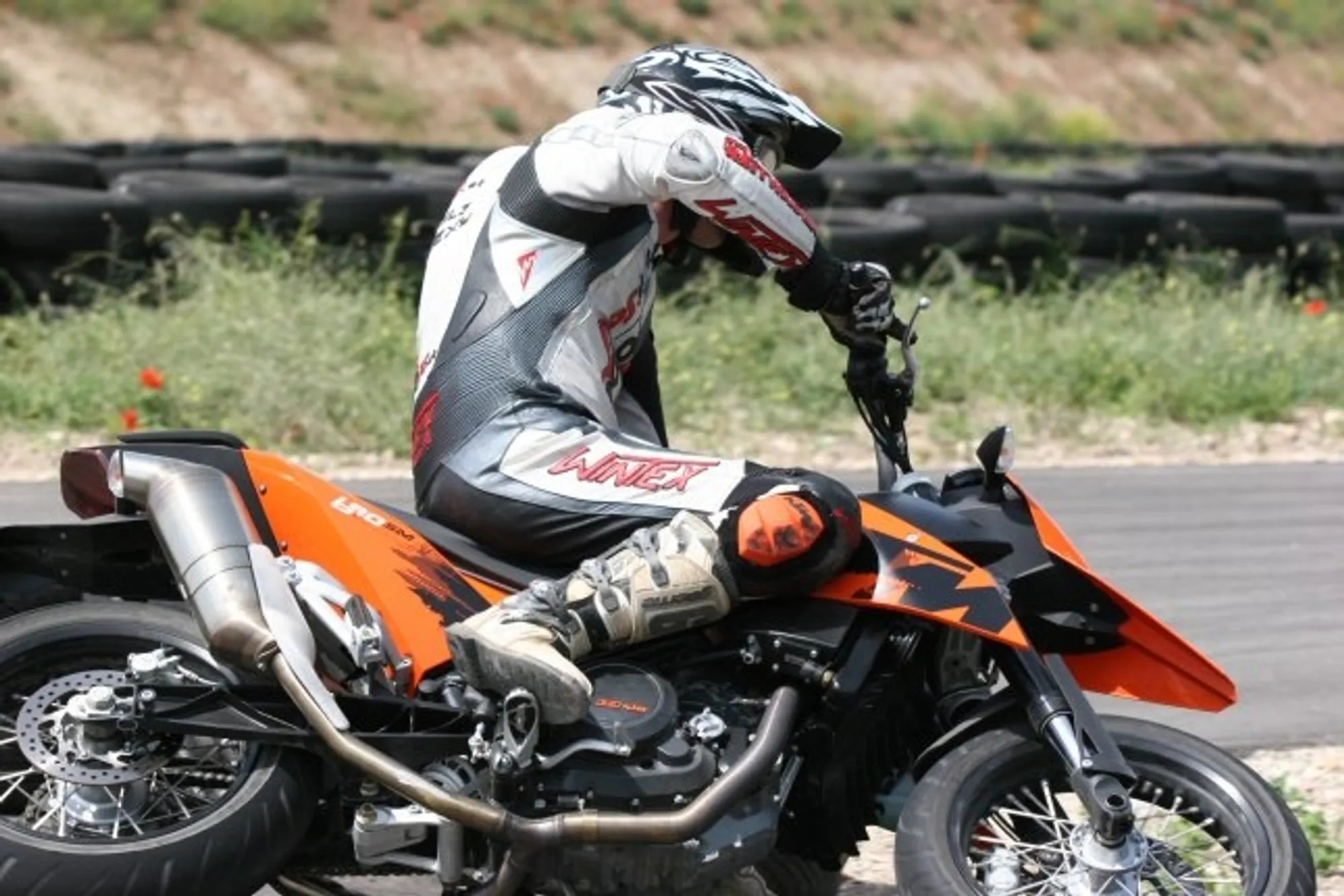 NastyNils riding a KTM 690 Supermoto at a supermoto track, leaning through a fast corner with aggressive body positioning. The orange and black bike is captured mid-turn on paved asphalt with tire barriers and green vegetation visible in the background. Full leathers and helmet worn, daylight conditions.