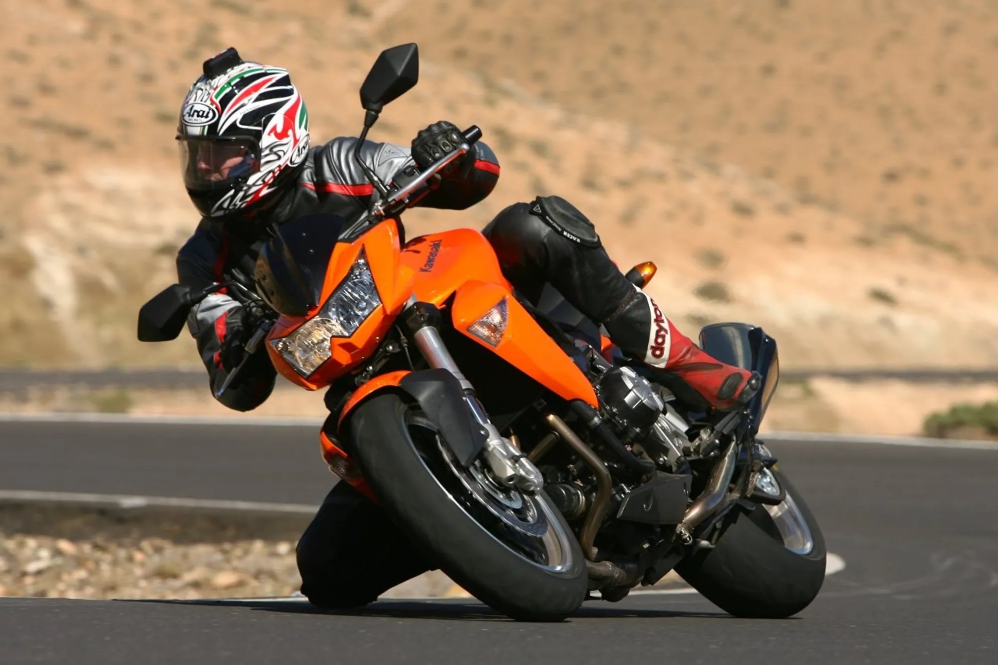 NastyNils riding a Kawasaki Z1000 in a kneedown on a winding road in the Canary Islands. The rider wears black leather gear and an Arai helmet with white and red graphics. Dry asphalt in daylight, arid desert landscape with sandy mountains visible in the background. The orange and black Z1000 is leaned to an extreme angle, demonstrating cornering capability and rider skill.