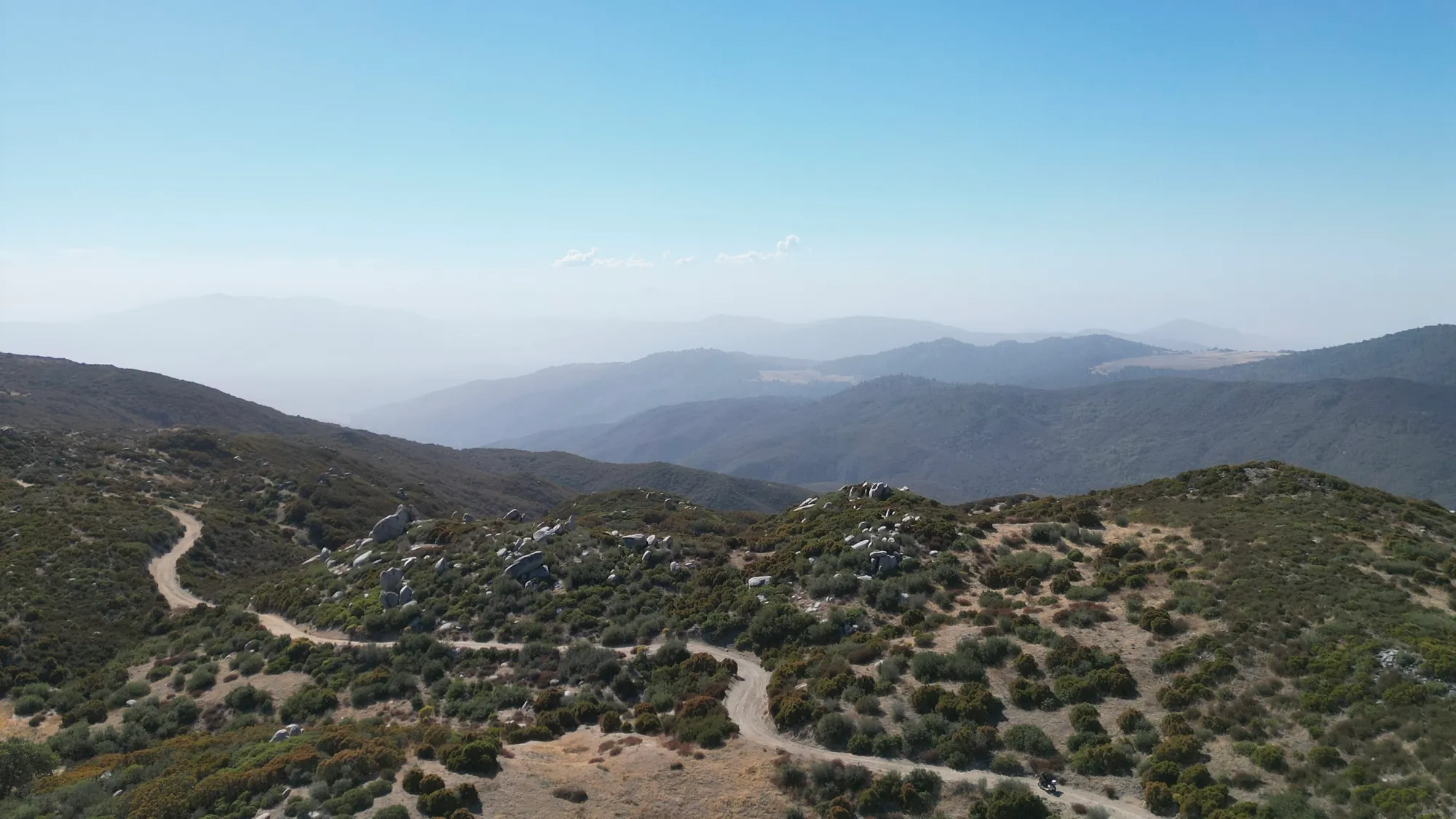Aerial drone view of Palomar Divide Road winding through chaparral-covered mountain ridges in San Diego County. Multiple S-curve sections descend through sparse vegetation with distant valley views visible in the haze. Gravel and packed-earth surface.