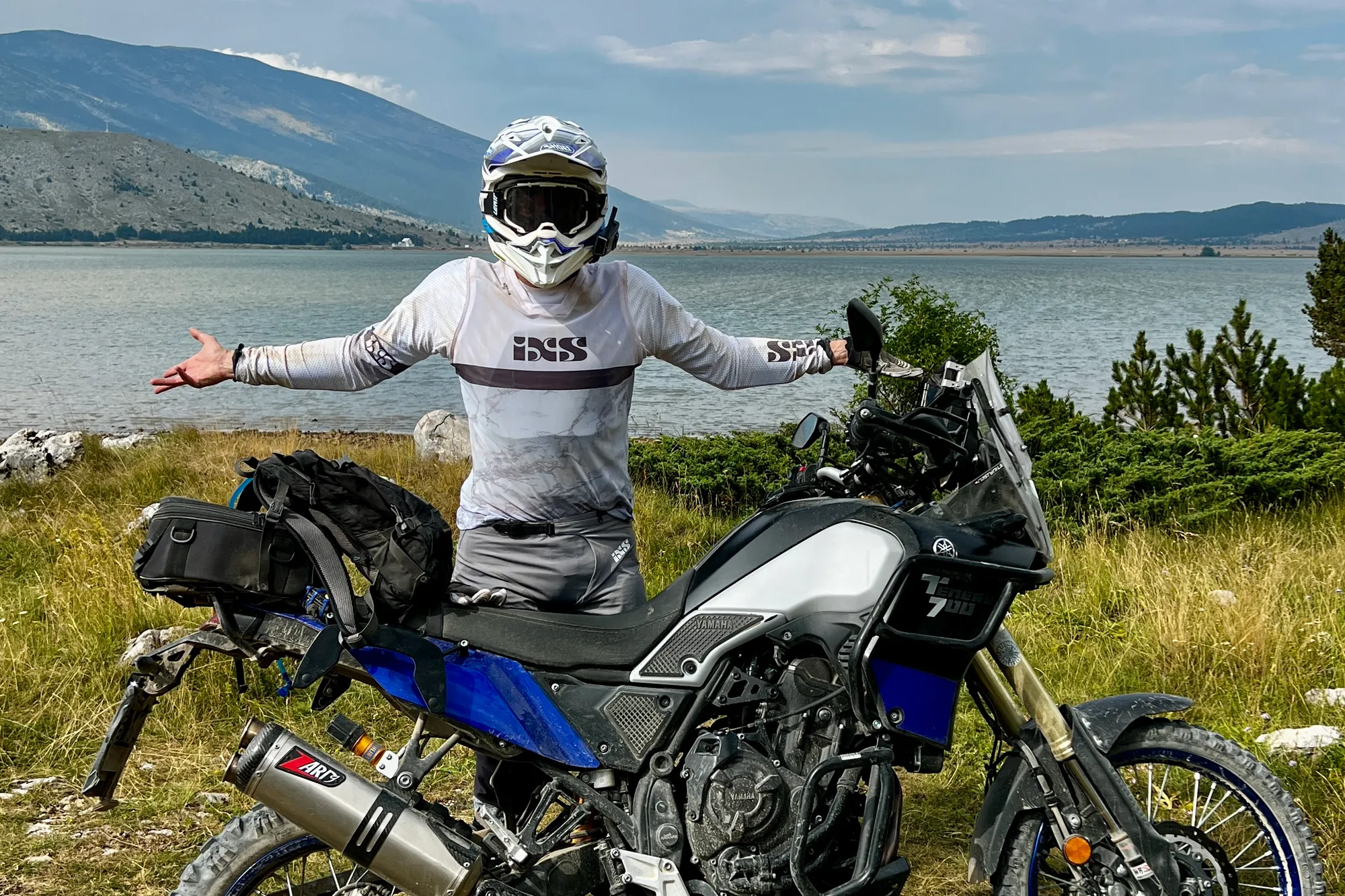 NastyNils stands beside a blue and white Yamaha Ténéré 700 adventure motorcycle at a lakeside location, arms spread in celebration. Mountains visible in the background across the water. The bike is loaded with travel gear and saddle bags, suggesting a long-distance adventure. Clear daylight, calm water, grassy terrain. NastyNils wears white IFS-branded protective gear and a full-face helmet.