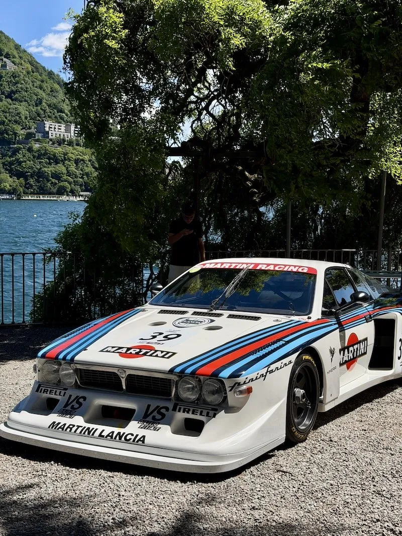 A Martini-liveried Lancia racing car on display at the Concorso d’Eleganza, the lake visible behind the trees.