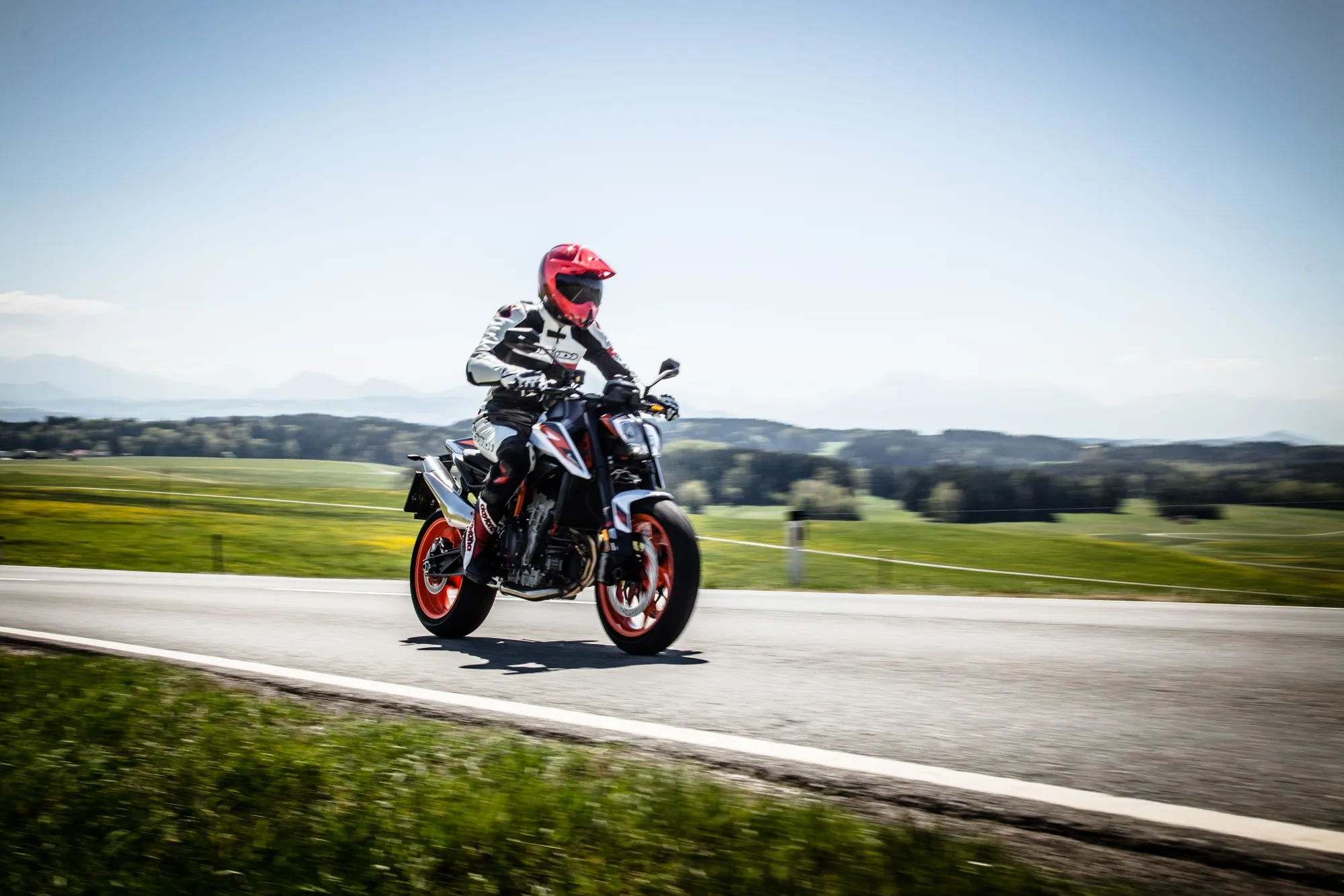 NastyNils riding a KTM 890 DUKE R on a rural country road in 2020. The bike is in motion across a straight section with rolling farmland in the background. NastyNils wears a pink helmet and white/black race suit, body position suggesting aggressive acceleration. The landscape shows green fields, clear blue sky, and distant hills. Dry asphalt, bright daylight conditions.