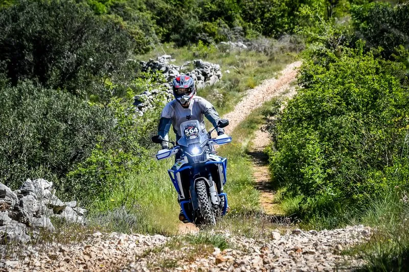 CFMOTO 450MT navigating a rocky off-road trail through Mediterranean scrub terrain with light-colored gravel and limestone, sparse vegetation, daylight.