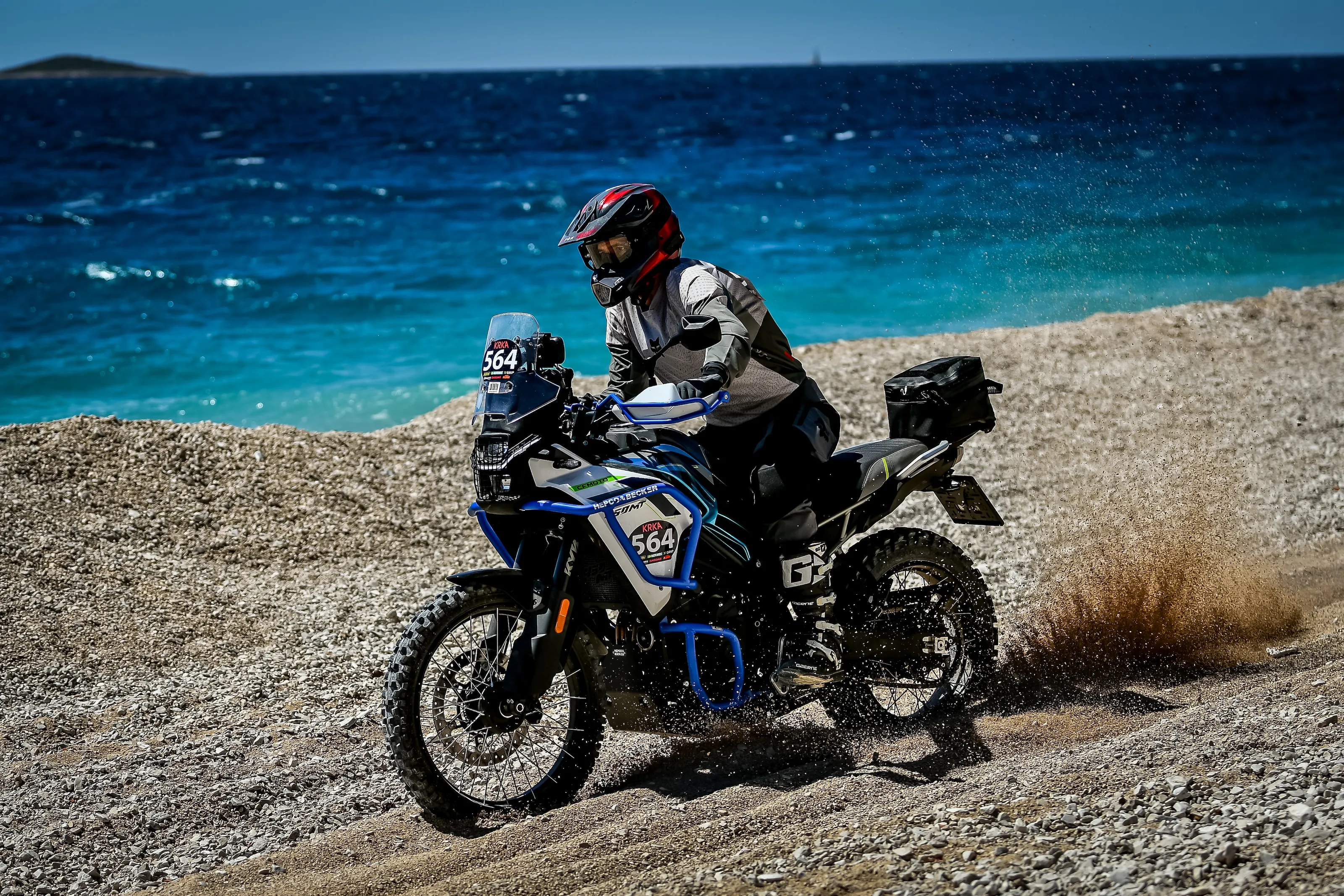 NastyNils riding a CFMOTO 450MT on a gravel coastal road in Croatia near Krka National Park with the turquoise Adriatic sea forming the scenic background.
