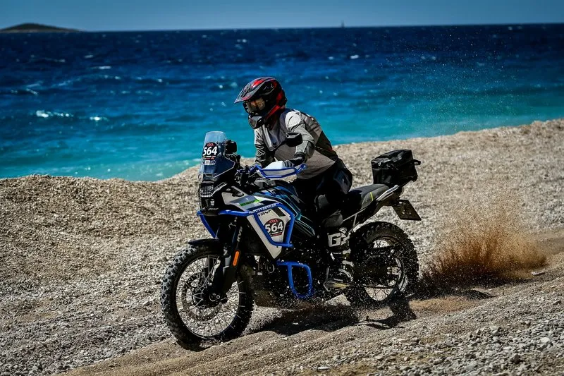 NastyNils riding a CFMOTO 450MT on a gravel coastal road in Croatia near Krka National Park with the turquoise Adriatic sea forming the scenic background.