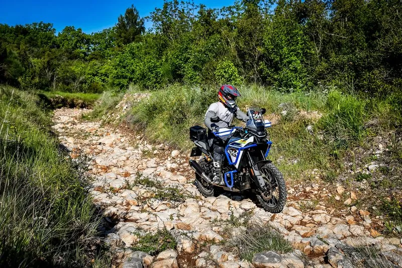 NastyNils on a CFMOTO 450MT adventure motorcycle on a rocky limestone trail with native Mediterranean vegetation, dynamic riding posture, rough gravel terrain in daylight conditions.