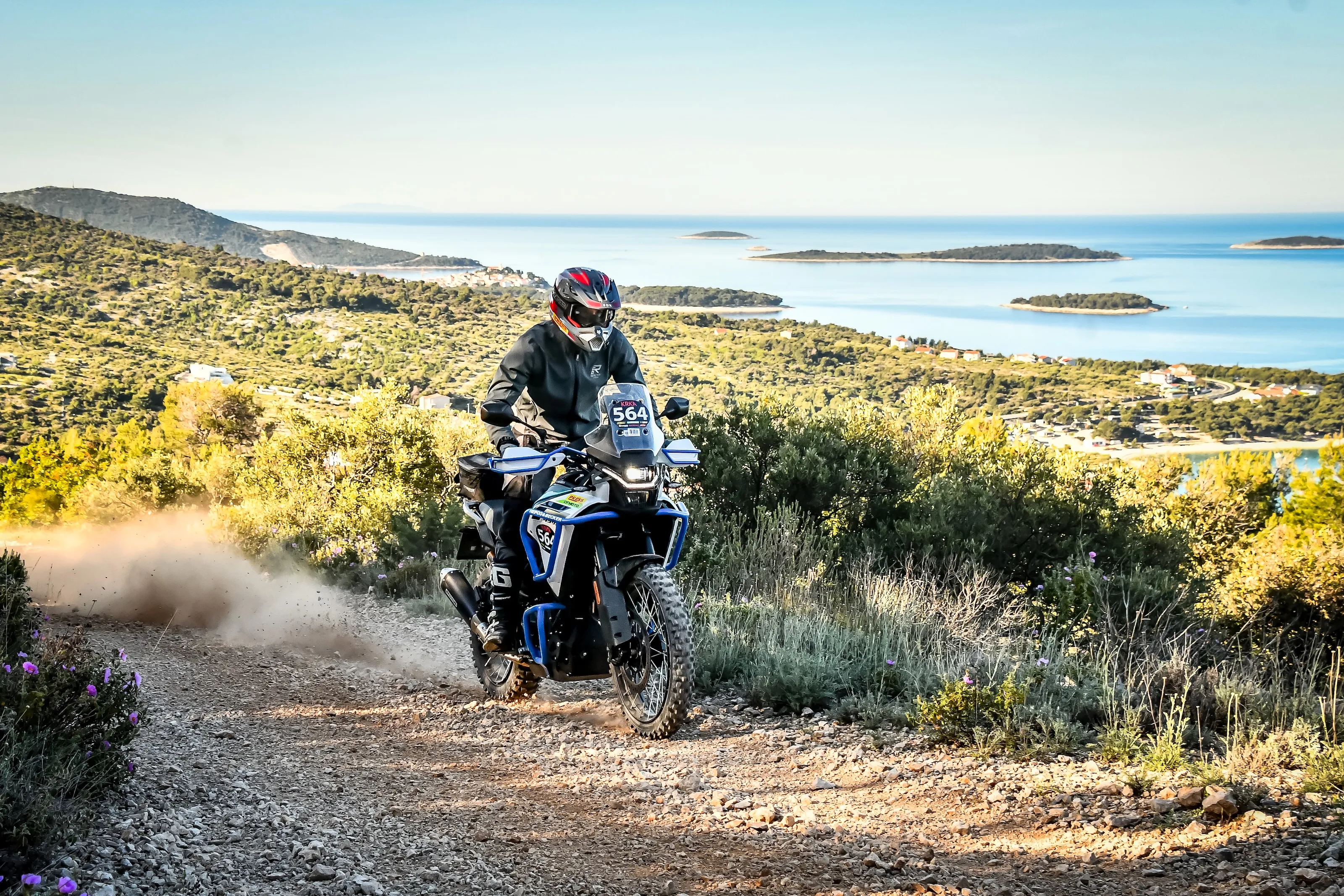 NastyNils riding a blue and white CFMOTO 450MT adventure motorcycle on rocky gravel terrain in a Mediterranean coastal region with islands and blue water visible in the distance, wearing rally bib 564.