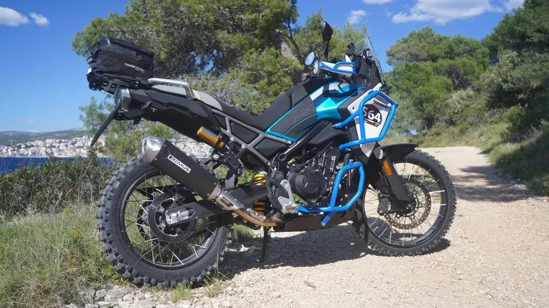 CFMOTO 450MT adventure motorcycle parked on a scenic coastal gravel road, equipped with luggage and a topcase, blue and black livery, Adriatic coast in the background.