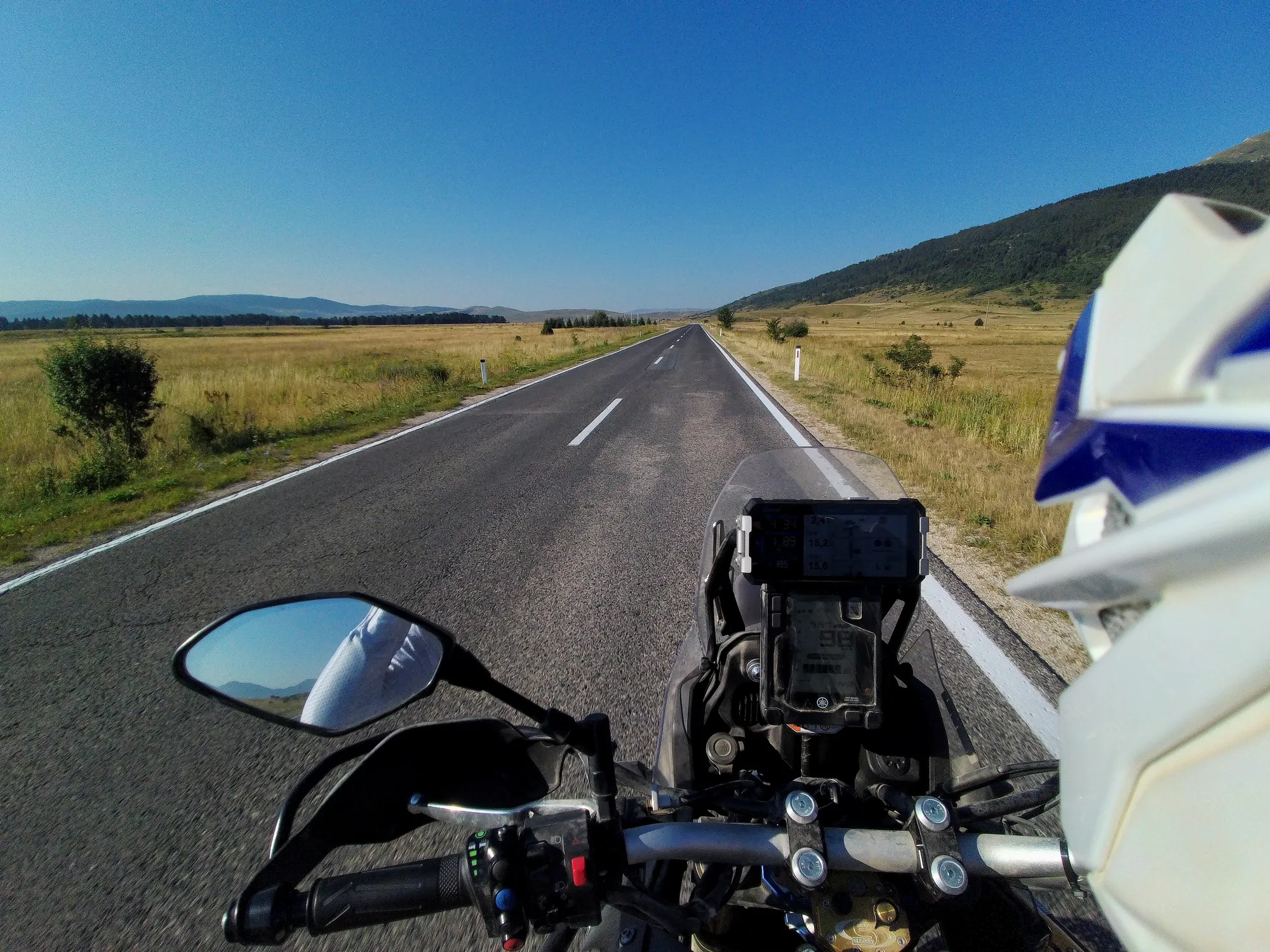 NastyNils rides a white-and-blue Yamaha Ténéré 700 down a straight paved road through East African savanna grassland. Mountains rise in the distance under clear blue sky. The landscape is arid and grassy with sparse vegetation. First-person perspective from the rider's viewpoint, showing handlebars, mirrors, and dashboard ahead on the dry asphalt road with white center markings.
