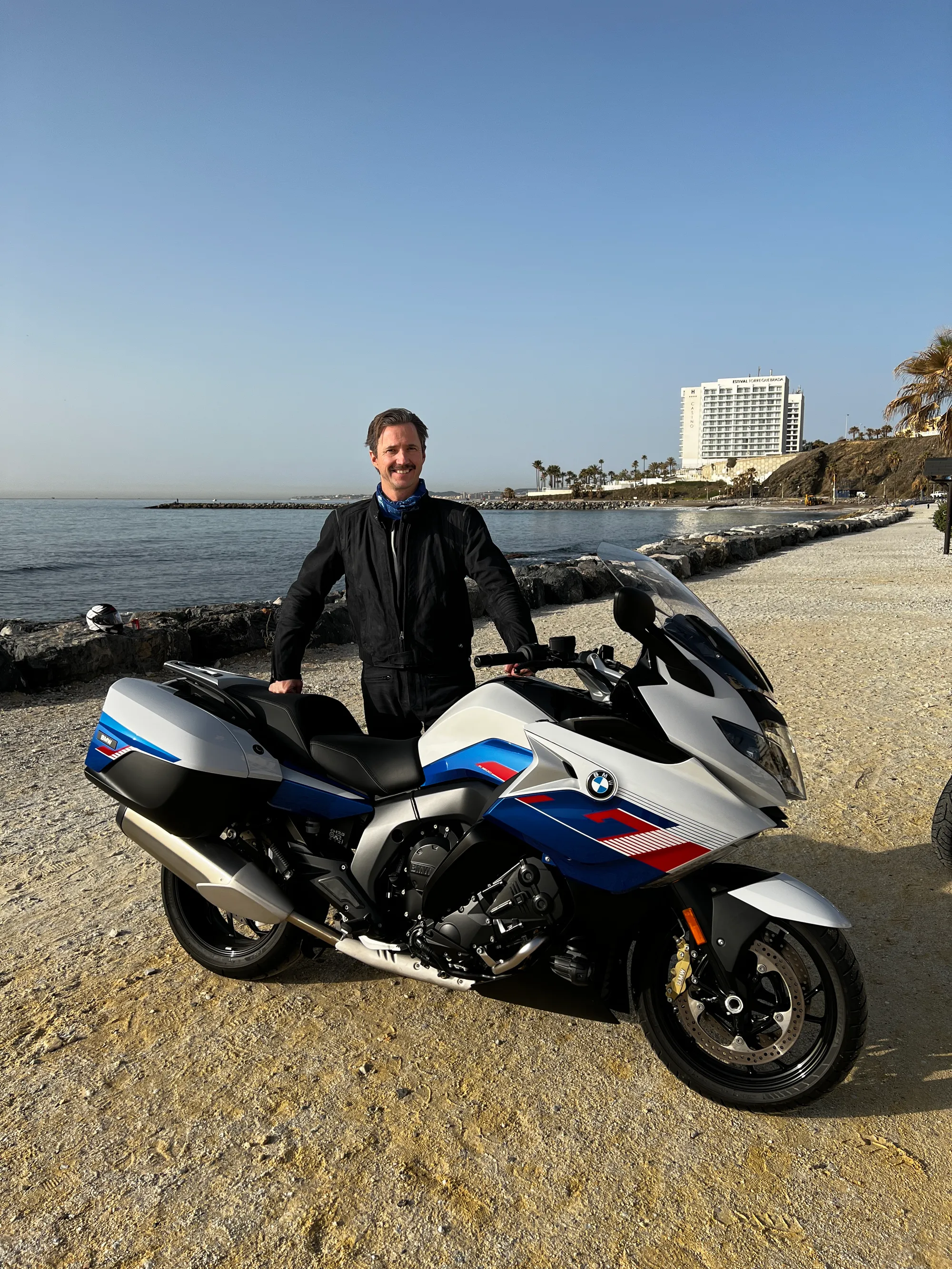 NastyNils and his BMW K1600GT on a quiet European street in Andalusia during a 2022 tour. The white, blue, and red touring bike sits parked against traditional stone architecture. Clear sunny day with no traffic. Nils poses casually in black leather next to the bike, demonstrating the K1600's compact footprint for urban navigation despite its touring capabilities and full fairing.