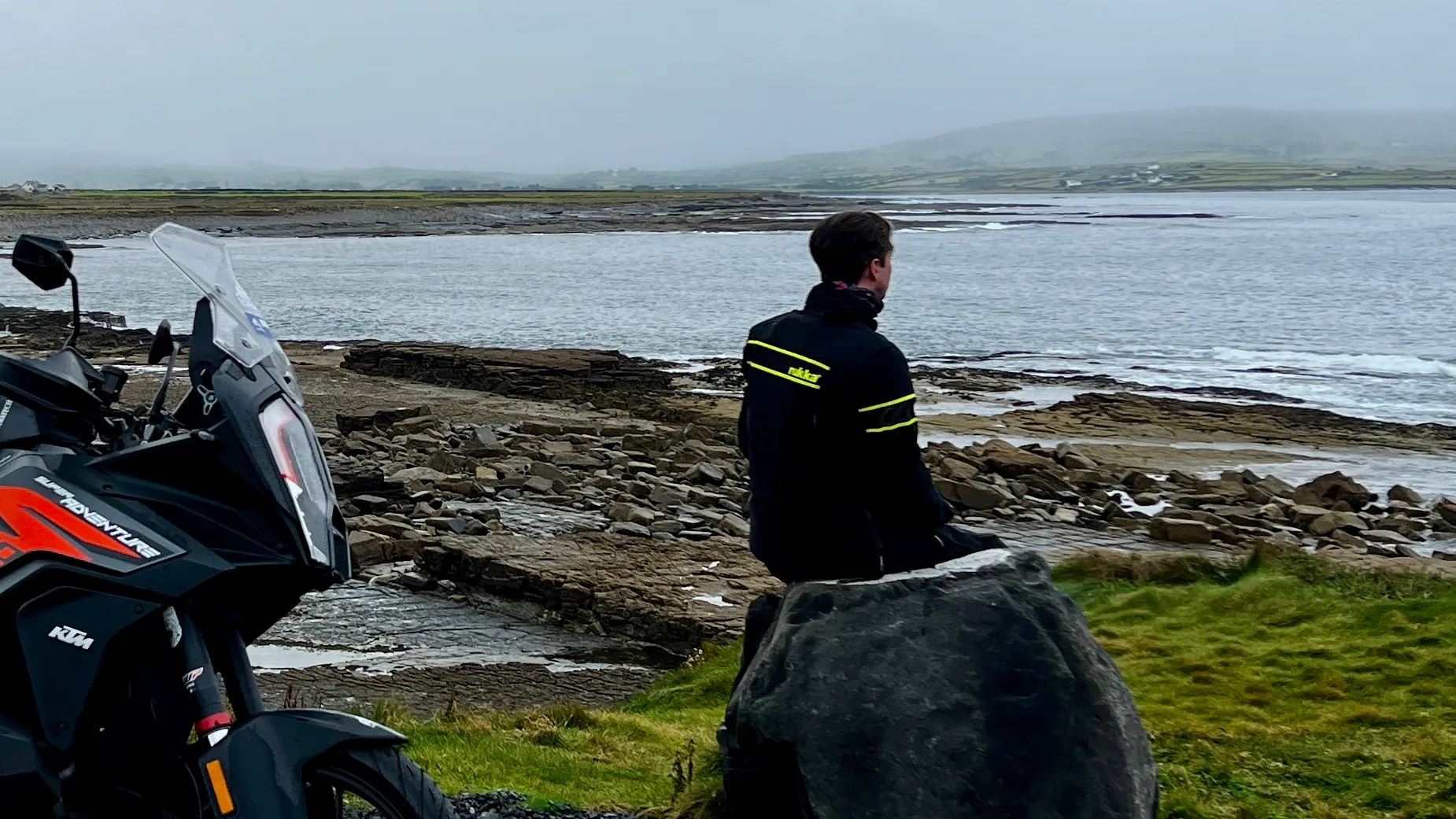 NastyNils stands beside his KTM 1290 Super Adventure S on a rocky coastal shore in Ireland, overlooking calm seas and distant fog-covered mountains. The bike's windscreen and protective fairing are visible. He wears a black riding jacket with reflective yellow stripes, gazing toward the horizon in a contemplative pose. Overcast weather, lush green grass visible in the foreground. The scene captures the essence of adventure touring in cool climate conditions.