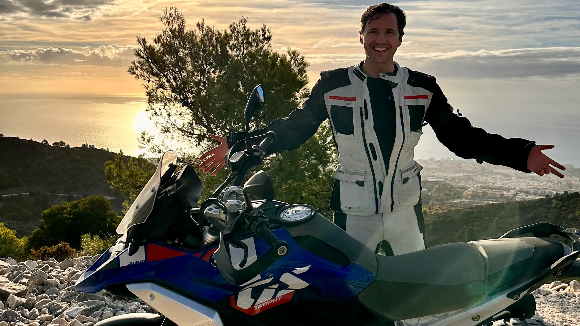 NastyNils posing with a BMW R 1300 GS adventure motorcycle at an Andalusian coastal overlook during golden hour. The bike sits on rocky terrain with Mediterranean vegetation and seascape visible in the background. NastyNils wears adventure touring gear with a white jacket and red accents, smiling with arms spread. The scene captures the joy and freedom of adventure-bike touring in southern Spain.