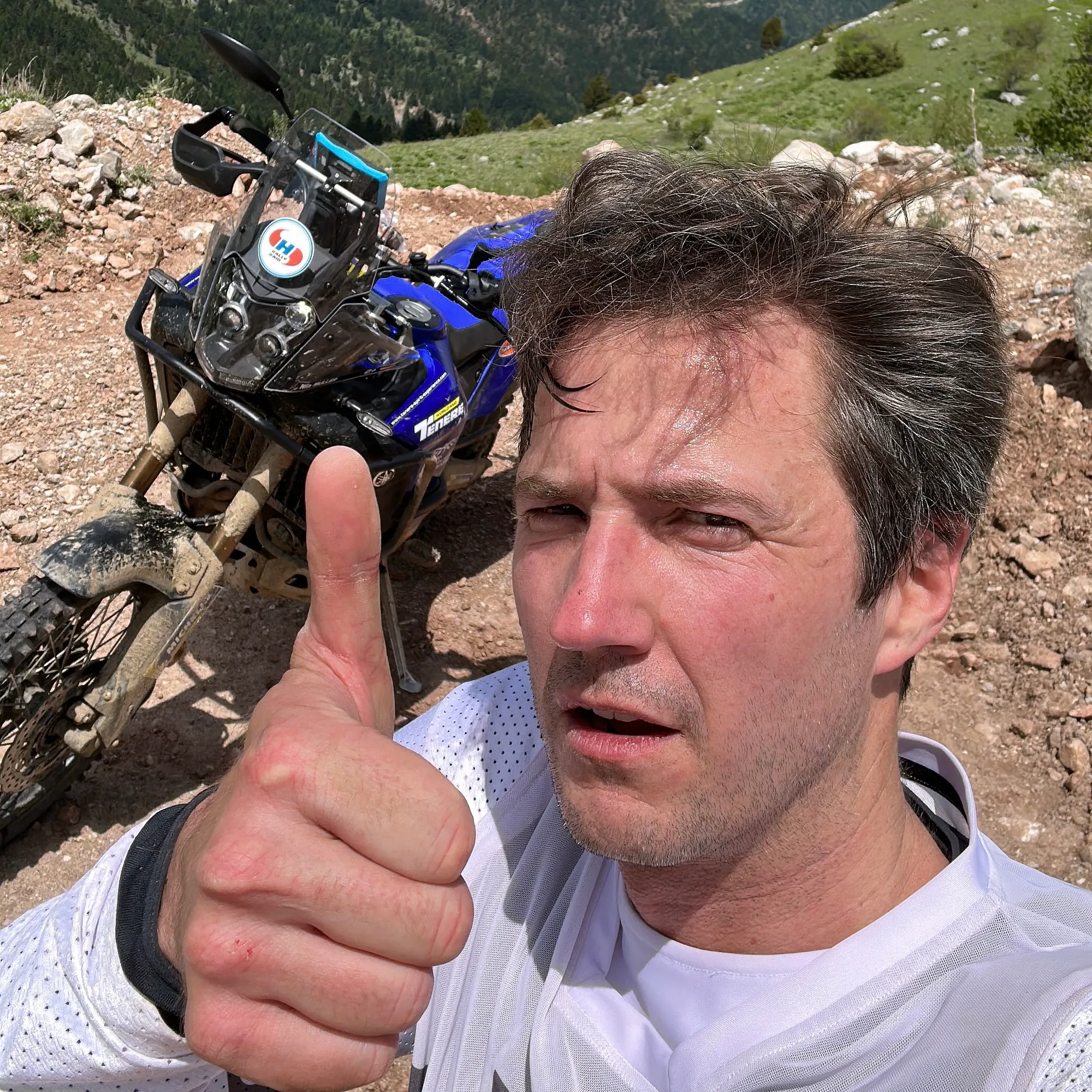 NastyNils celebrates a successful adventure ride on his Yamaha Tenere 700 in high-altitude mountain terrain. Rocky ground with sparse vegetation and distant peaks visible. Nils wears a white t-shirt and gives a thumbs-up to the camera, expressing satisfaction with the bike's hard-enduro performance. The Tenere 700 is parked in the background on loose rocky ground.