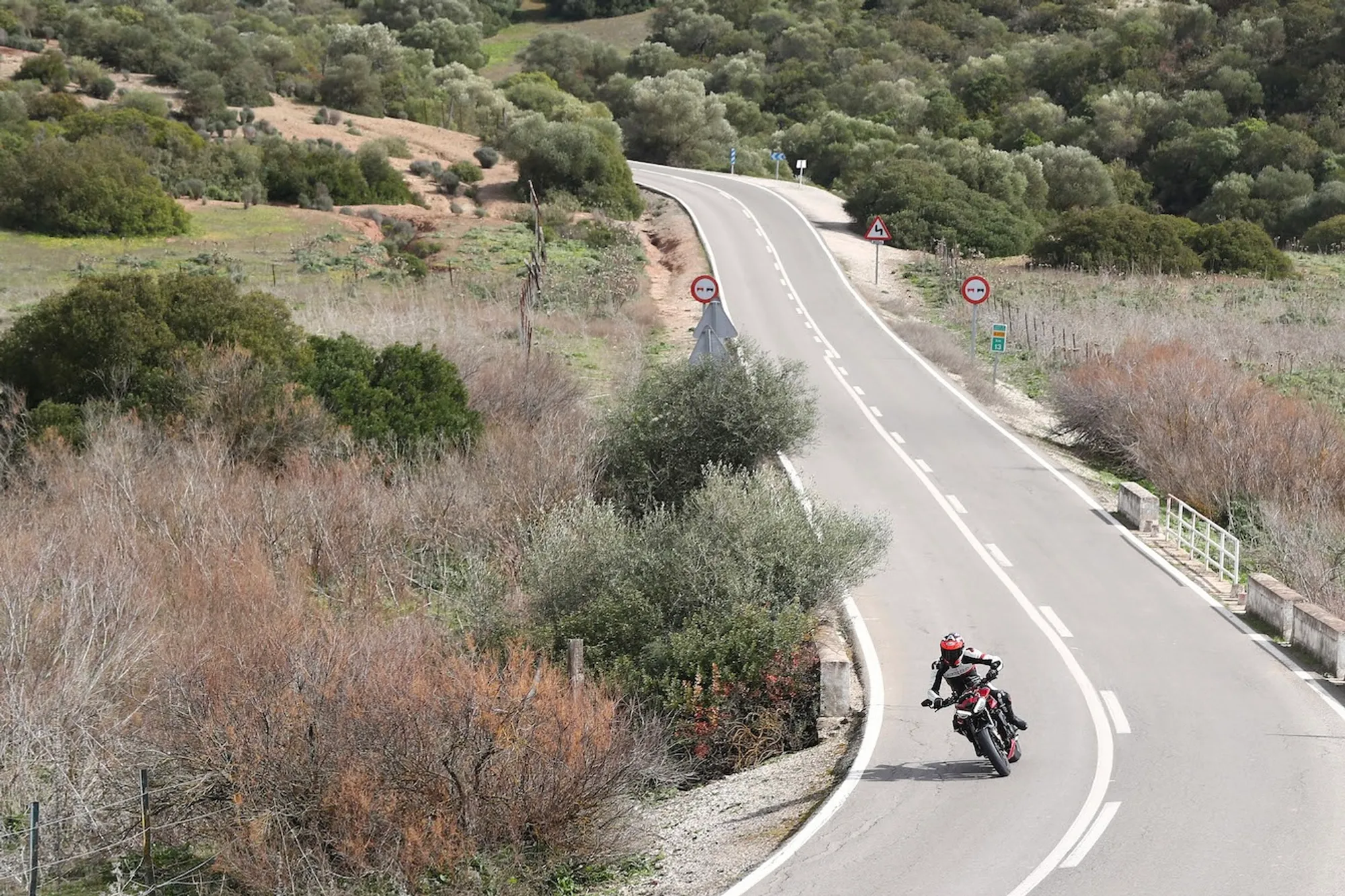 NastyNils riding a Triumph Street Triple RS through a winding Mediterranean country road, leaning deeply through a sweeping left-hander. Rolling hills with sparse olive trees and scrub vegetation frame the scene. Clear daylight, dry asphalt, rider in black leathers and full-face helmet. Guardrails and road signage visible ahead on the rising curve.