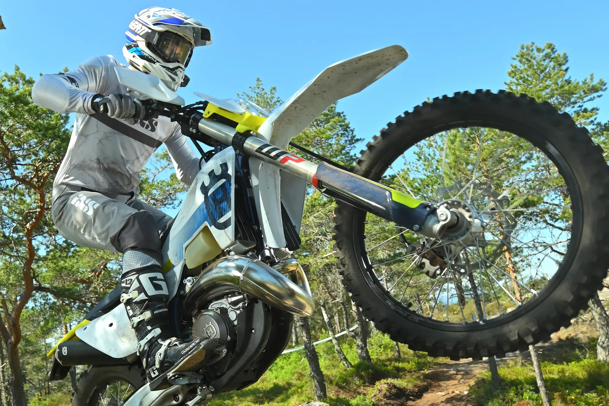 NastyNils rides a Husqvarna TE300 on a sandy off-road trail through forest, performing a bunny-hop or wheelie maneuver. Full enduro gear visible: white chest protector, protective helmet, aggressive riding posture. The two-stroke single-cylinder enduro machine's trellis frame and lightweight design are evident. Daylight, clear skies, natural forest setting.