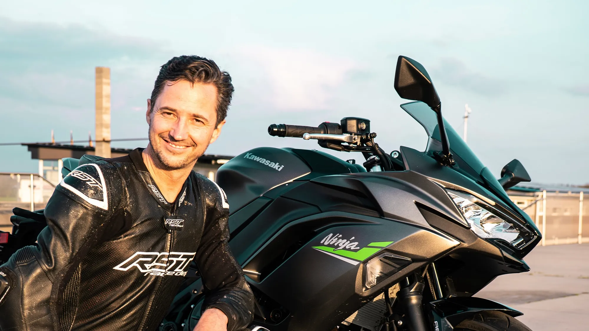 NastyNils with a black-and-green Kawasaki Ninja ZX6R on an urban rooftop. The rider wears black leather gear with Kawasaki and RAZE branding. The supersport bike displays its characteristic aggressive LED headlight design and fairings. Industrial buildings and overcast sky visible in the background. Posed presentation shot in daytime natural light.