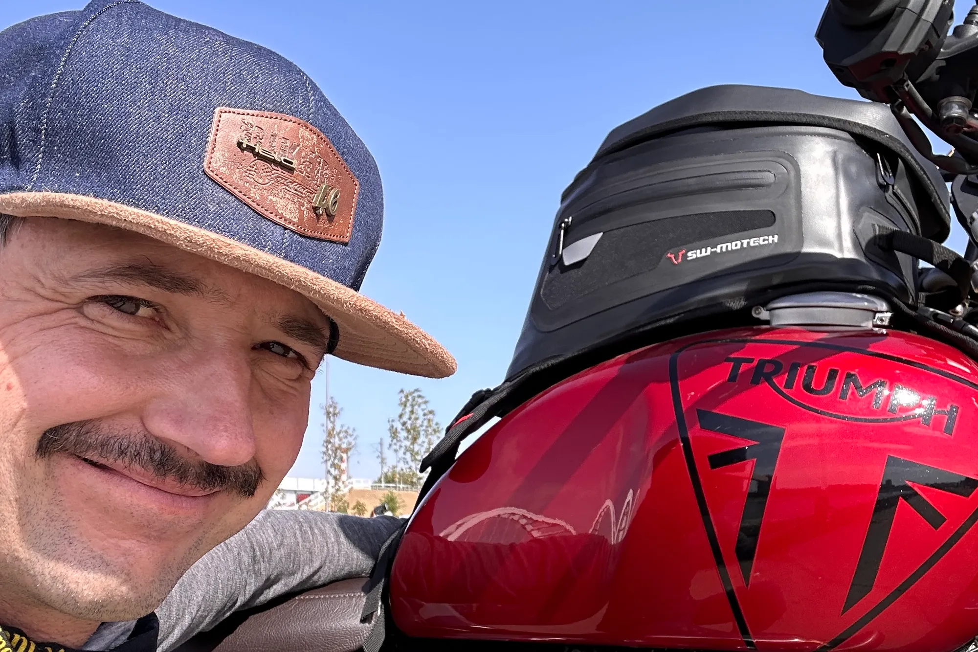 NastyNils poses for a selfie next to a red Triumph classic motorcycle in a mountainous, sparse landscape. He wears a denim-style cap and dark jacket with neutral-toned gear. The Triumph fuel tank is prominently visible with its distinctive red finish and classic styling. Background shows rocky, hilly terrain with sparse vegetation. Daylight, clear blue sky.