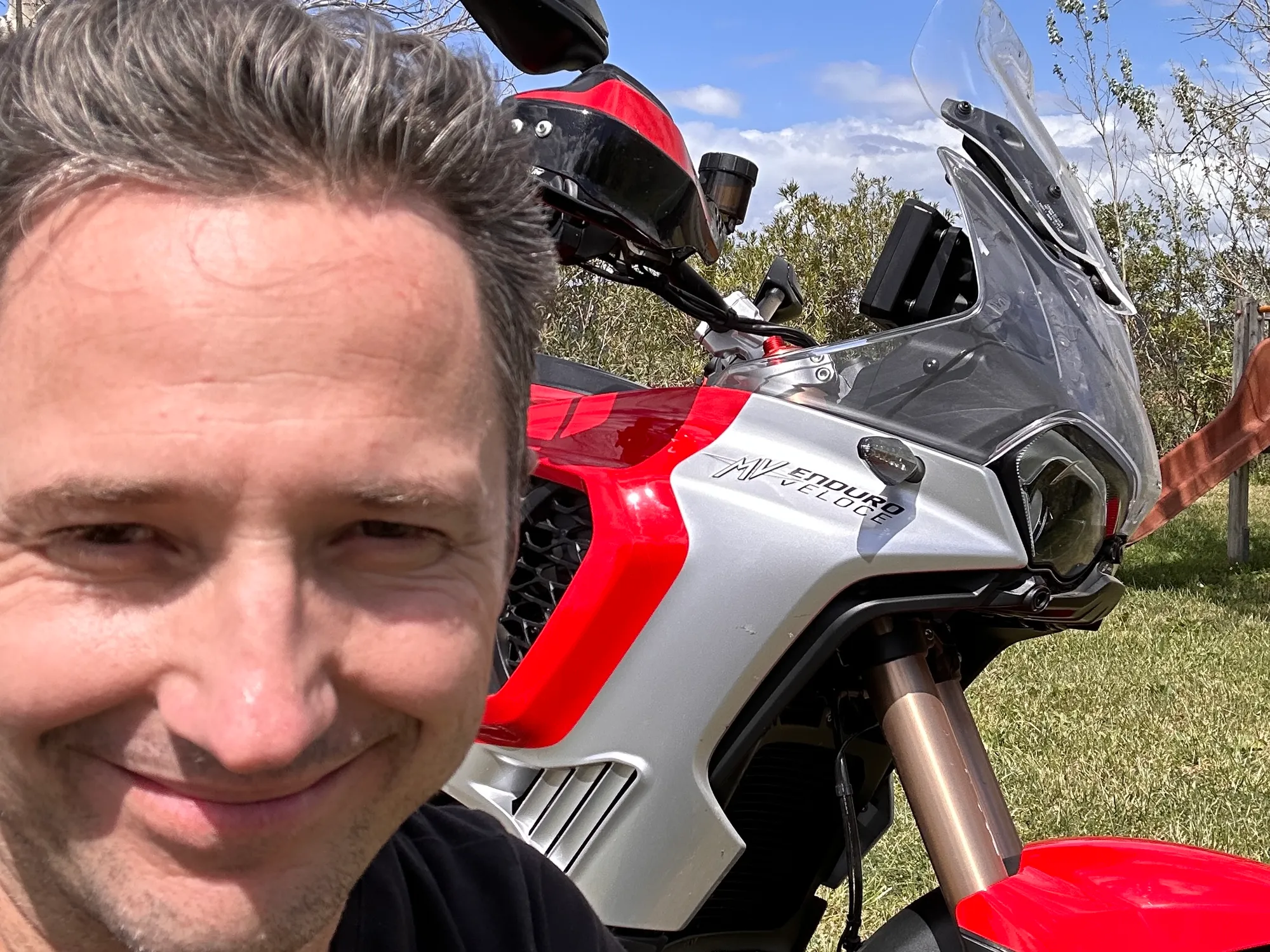 NastyNils poses with an MV Agusta Enduro Veloce MY2024 in a rural, tree-lined setting. He smiles at the camera in the foreground; the red-and-white sport-touring bike sits behind him at a 3/4 angle. Daylight, pastoral background. The bike's distinctive sport-tourer fairing and MV Agusta branding are clearly visible.