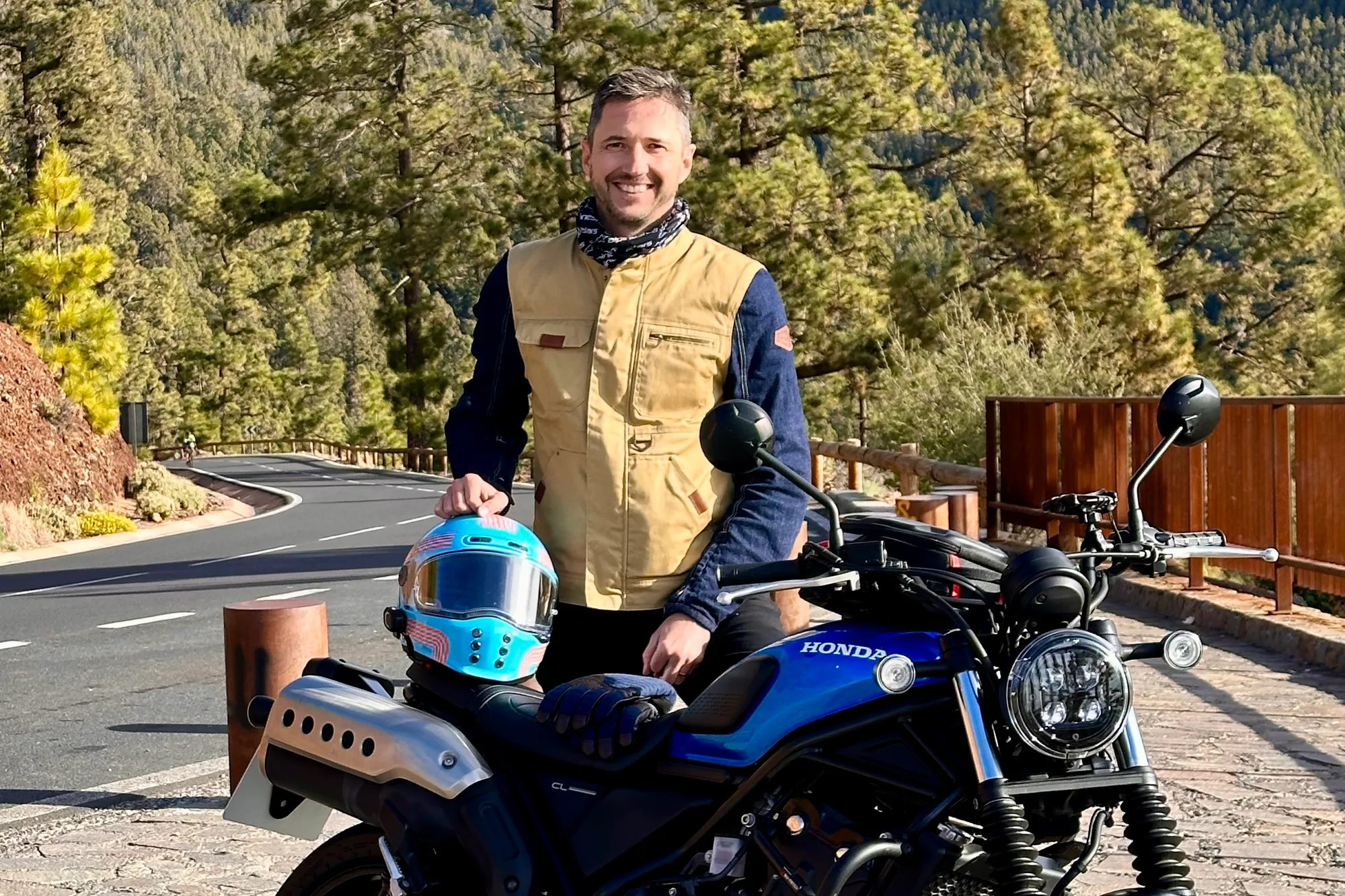 NastyNils poses with a Honda CL500 cruiser on a mountain road surrounded by forest. The bike is parked, facing slightly right, with a winding asphalt road and dense tree-covered landscape in the background. NastyNils wears a tan/beige vest over a dark blue long-sleeve shirt and holds a blue full-face helmet. Daylight, clear weather, scenic mountain setting. The CL500's retro-cruiser styling and lightweight frame are clearly visible.