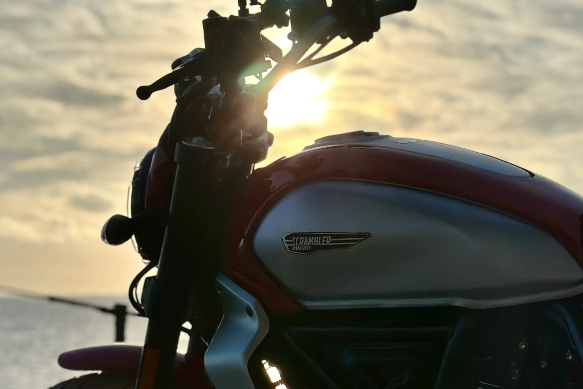 A Ducati Scrambler MY2023 parked overlooking the ocean at golden hour in the Canary Islands. The motorcycle is photographed in three-quarter view with the sun behind the bike, creating dramatic backlighting. The water and horizon are visible in the soft background. No rider present. High-quality composition with natural lighting.