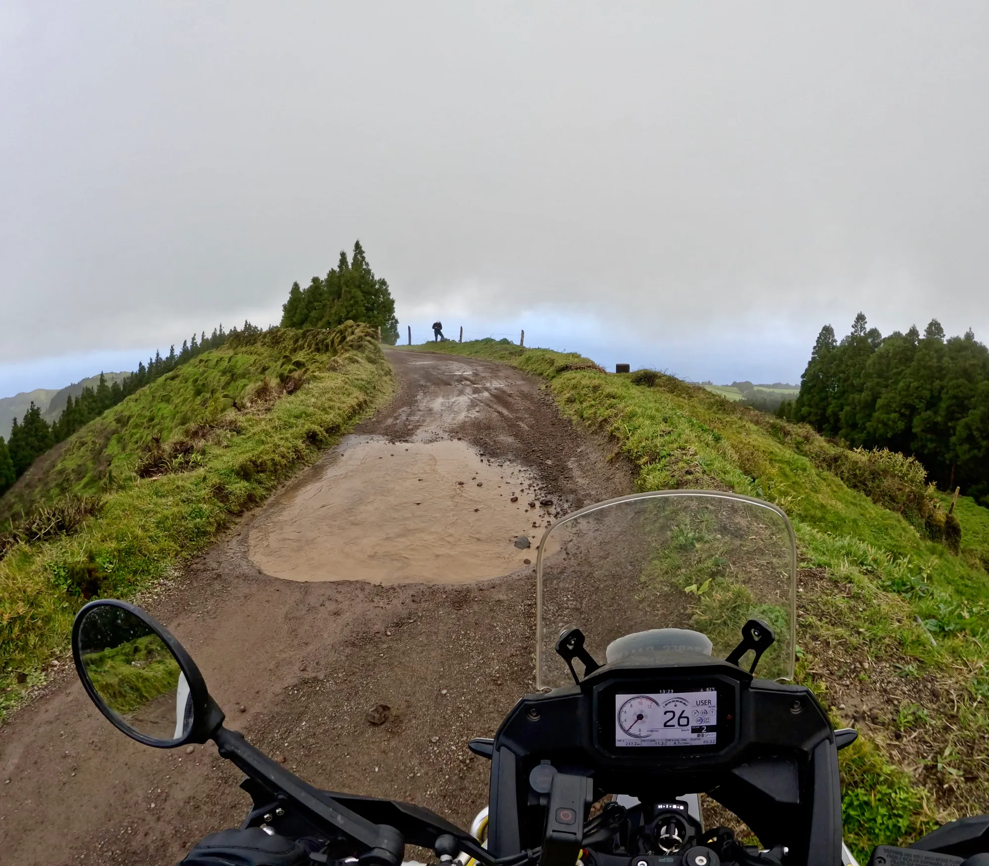 POV shot from NastyNils riding a Honda Transalp 700 up a steep dirt and gravel mountain trail. The adventure bike's digital dashboard and windscreen visible in the foreground. Alpine terrain with green hillsides, exposed soil track, dense forest on both sides, and a figure visible on the distant ridge line. Overcast mountain weather, steep climbing gradient, single-track off-road trail.