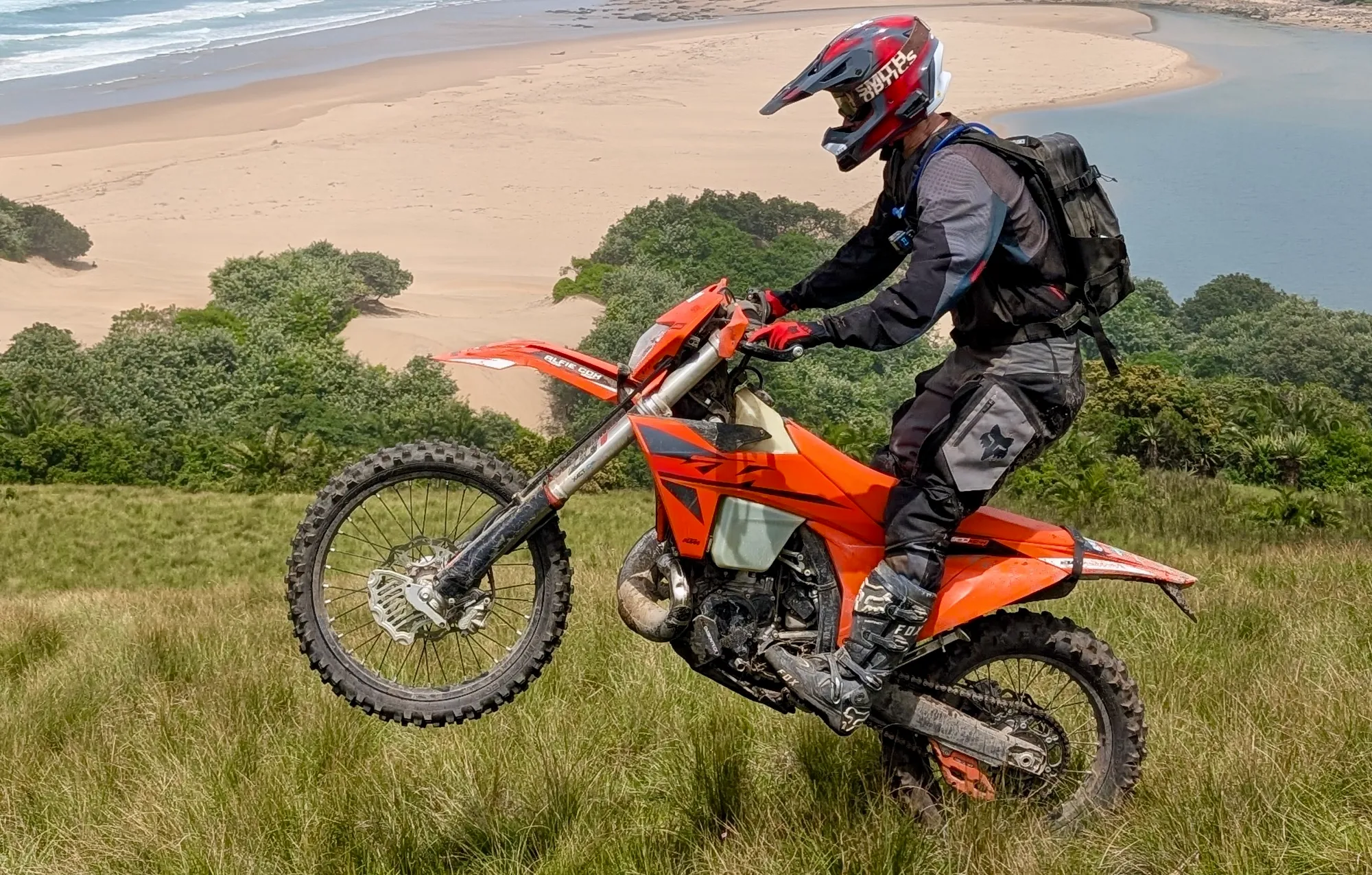 NastyNils performing a wheelie on a KTM 300 EXC Enduro in a coastal meadow in South Africa (February 2025). Beach and ocean visible in distant background, green grassland in foreground. Rider in full protective gear including helmet and body armor, standing on pegs. Natural off-road terrain, technical riding demonstration.