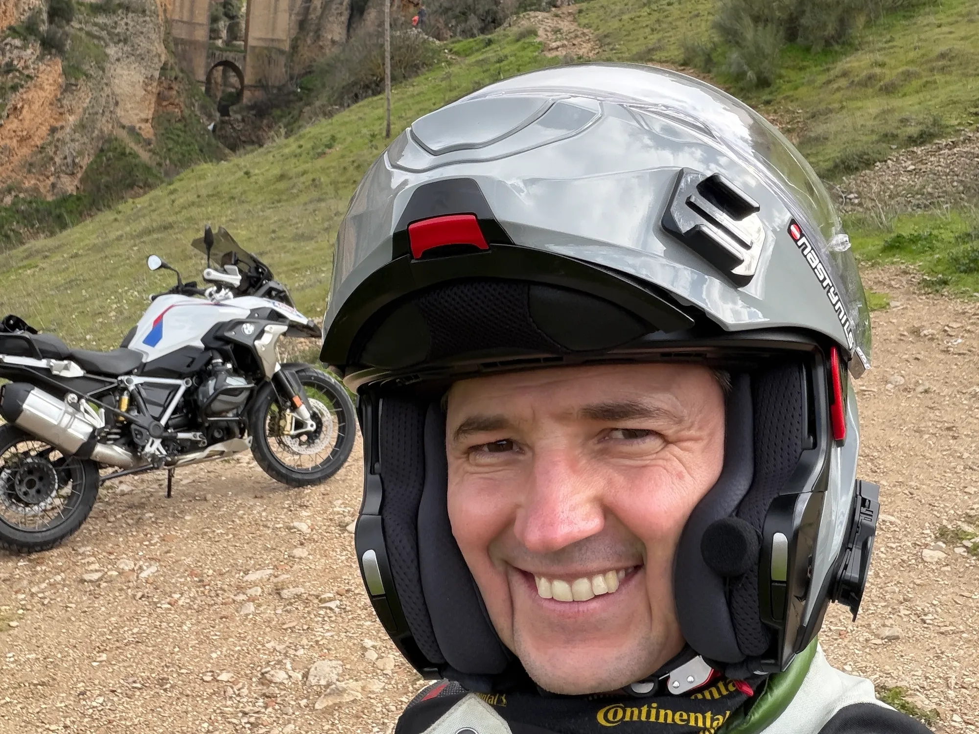 NastyNils in portrait, wearing a white Continential-branded full-face helmet on a gravel road in Andalusia. His BMW R1250GS adventure bike is parked behind him on the left. Rolling green hills visible in background. Sunny, dry daylight conditions. Nils is smiling directly at camera, dressed in adventure riding gear suitable for off-road terrain.