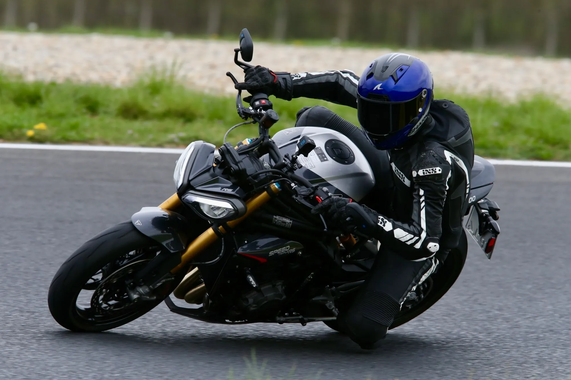NastyNils riding a Triumph Speed Triple RS on a racetrack, leaning aggressively through a high-speed corner in full black racing leather with white stripes and a blue helmet. The bike displays the characteristic white and black Triumph color scheme. Racetrack asphalt with gravel trap and safety guard rail visible in background.