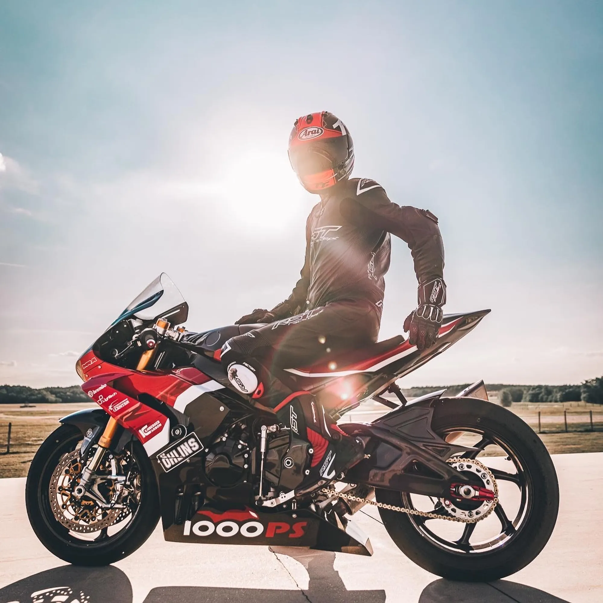 NastyNils performing a wheelie stunt on a Honda CBR1000RR Fireblade MY2020 in a track-day setting. The rider wears a black leather racing suit with red and grey Arai helmet. The bike's striking red and black livery with visible 1000PS branding contrasts against the flat concrete paddock and open sky. Bright daylight, high-resolution action photography capturing track-day prowess.
