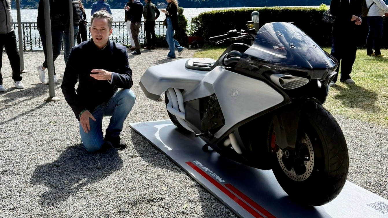 Nils kneels next to the BMW Motorrad Vision K18 concept on its display platform at the Concorso d'Eleganza, with Lake Como in the background.