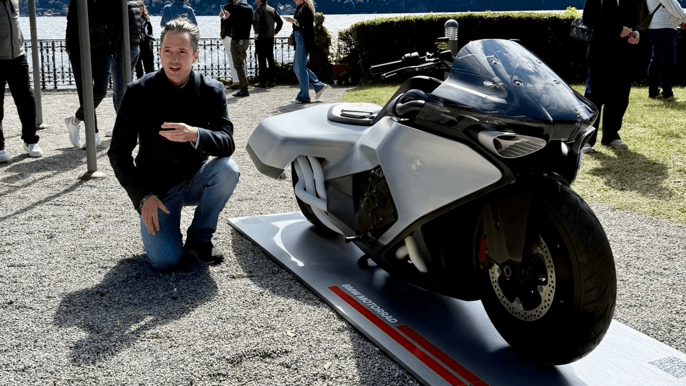 Nils kneels next to the BMW Motorrad Vision K18 concept on its display platform at the Concorso d'Eleganza, with Lake Como in the background.