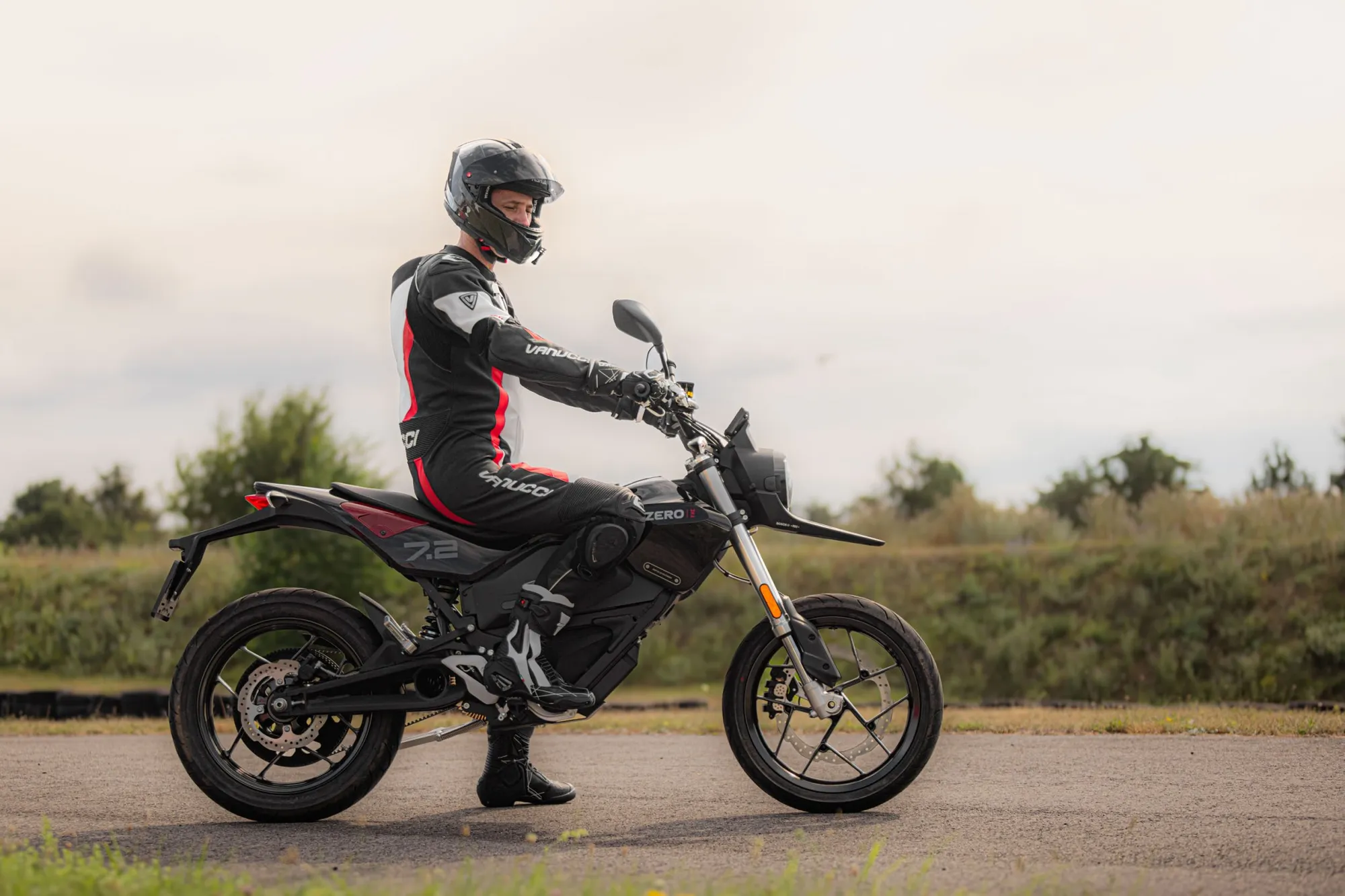NastyNils seated on a Zero FXE electric motorcycle in a static, posed shot on a gravel road surrounded by open fields. The rider wears black and red racing leathers with a full-face helmet. The FXE is a compact naked-style electric bike with minimal bodywork and a visible Zero badge. Overcast daylight, dry surface, rural landscape in soft focus behind.