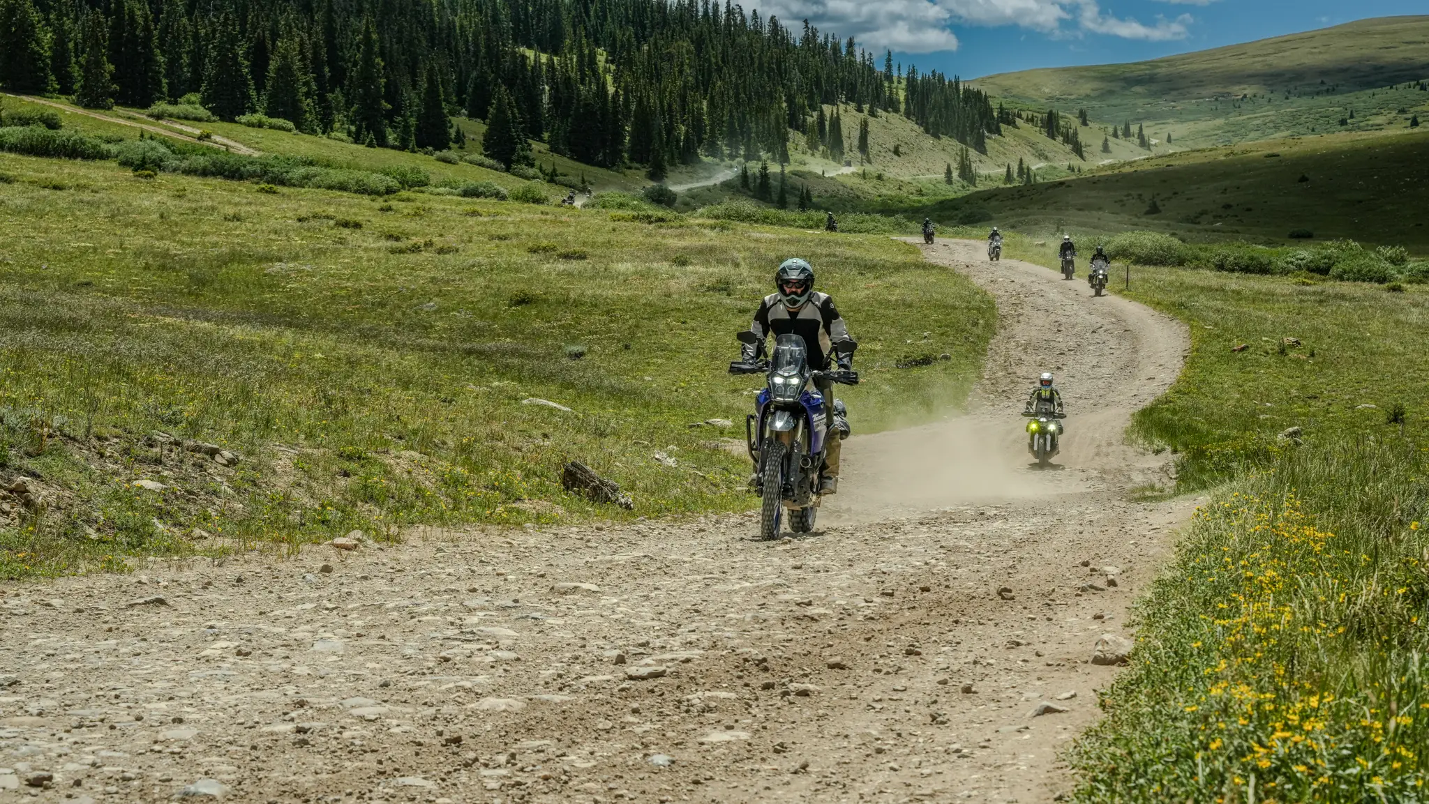 A convoy of adventure riders descending a winding gravel trail through an open Colorado alpine meadow, pine-covered slopes and rolling hills behind.