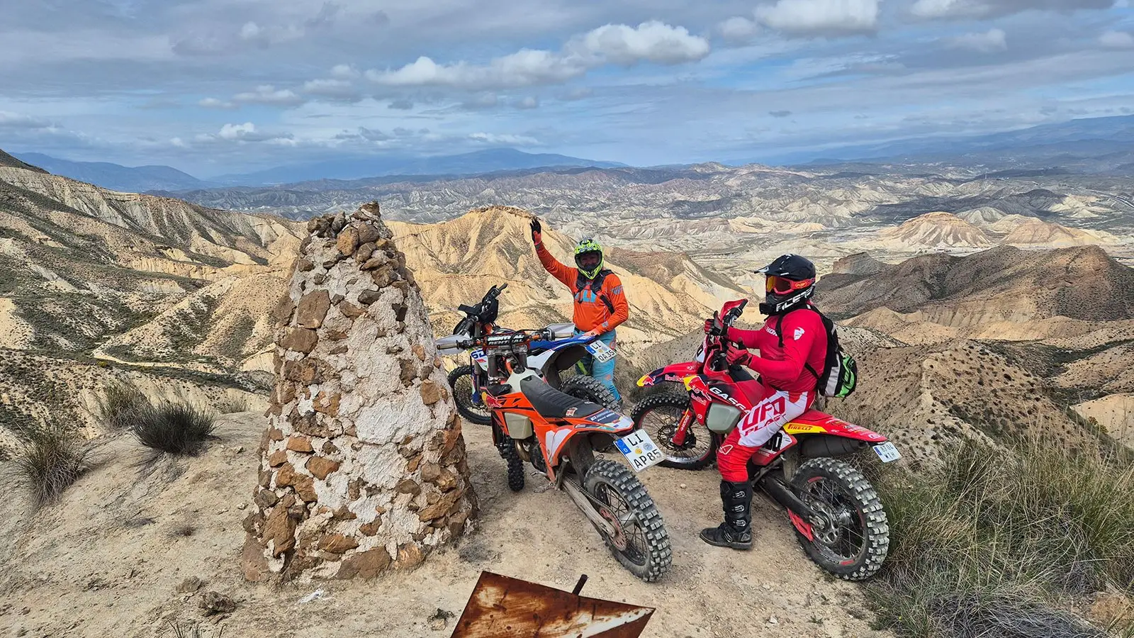 Two enduro riders with their bikes beside a stone cairn on a ridge above badlands in Andalusia.