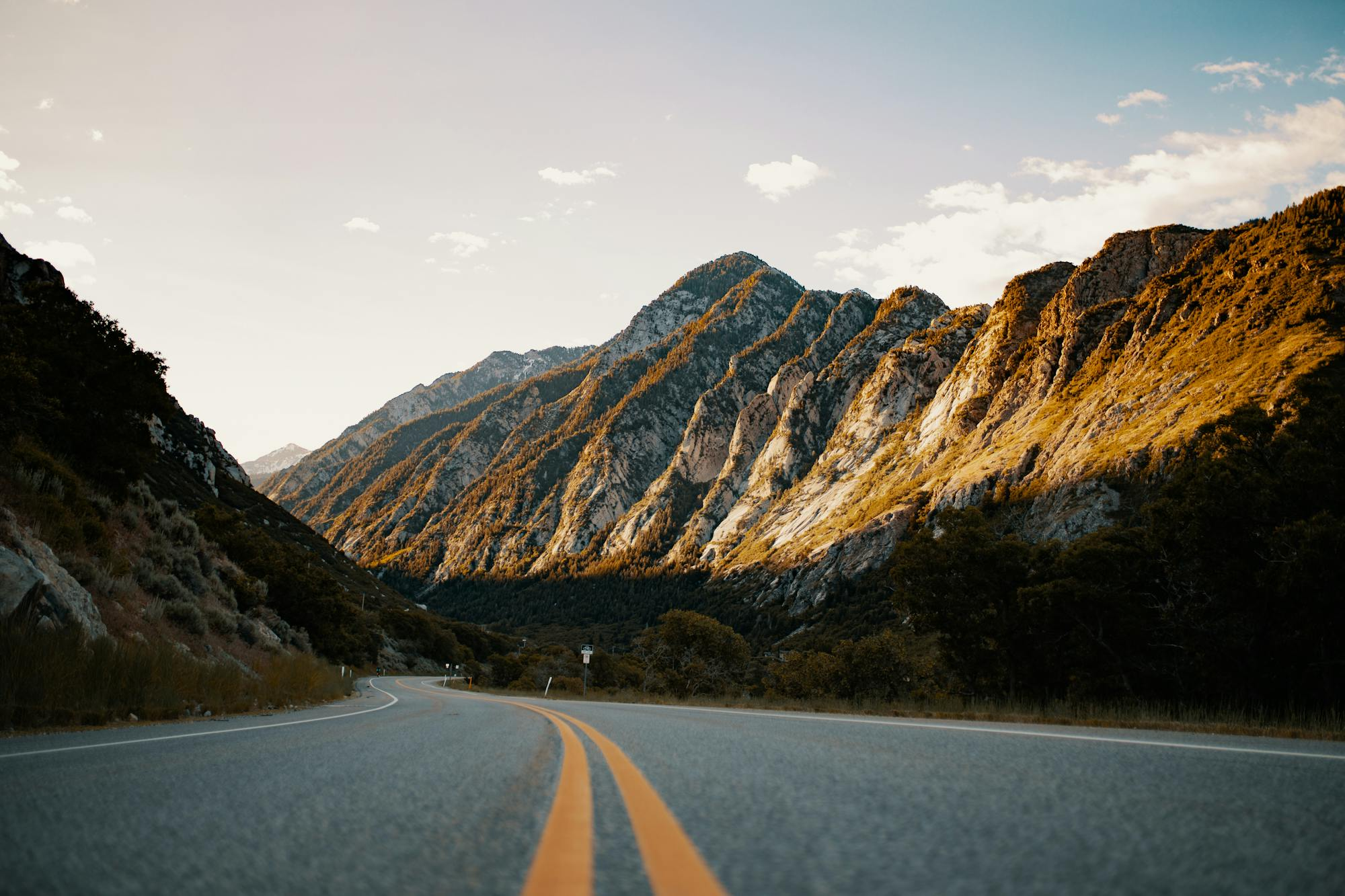 A scenic view of Angeles Crest Highway winding through rugged Southern California canyon terrain. Rocky mountainsides with golden earth tones frame the asphalt road with tight sweeping curves. Double yellow center line visible, sparse vegetation along the shoulders, clear blue sky with white clouds. Daylight, dry conditions. No motorcycle or rider visible. Iconic location for canyon-road enthusiasts.