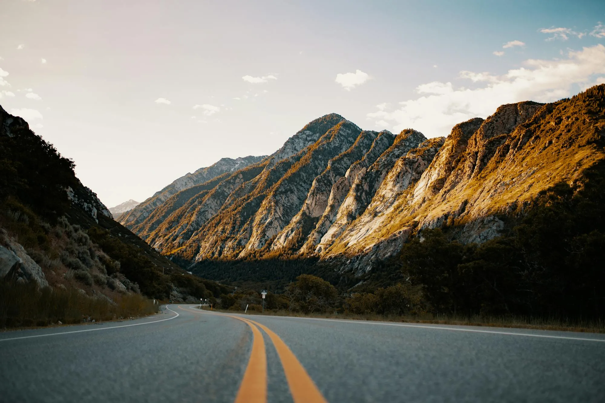 A scenic view of Angeles Crest Highway winding through rugged Southern California canyon terrain. Rocky mountainsides with golden earth tones frame the asphalt road with tight sweeping curves. Double yellow center line visible, sparse vegetation along the shoulders, clear blue sky with white clouds. Daylight, dry conditions. Iconic location for canyon-road enthusiasts.