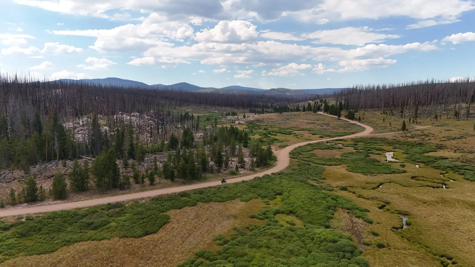 Aerial view of a dirt road winding through a green Colorado valley with burnt forest and mountains beyond