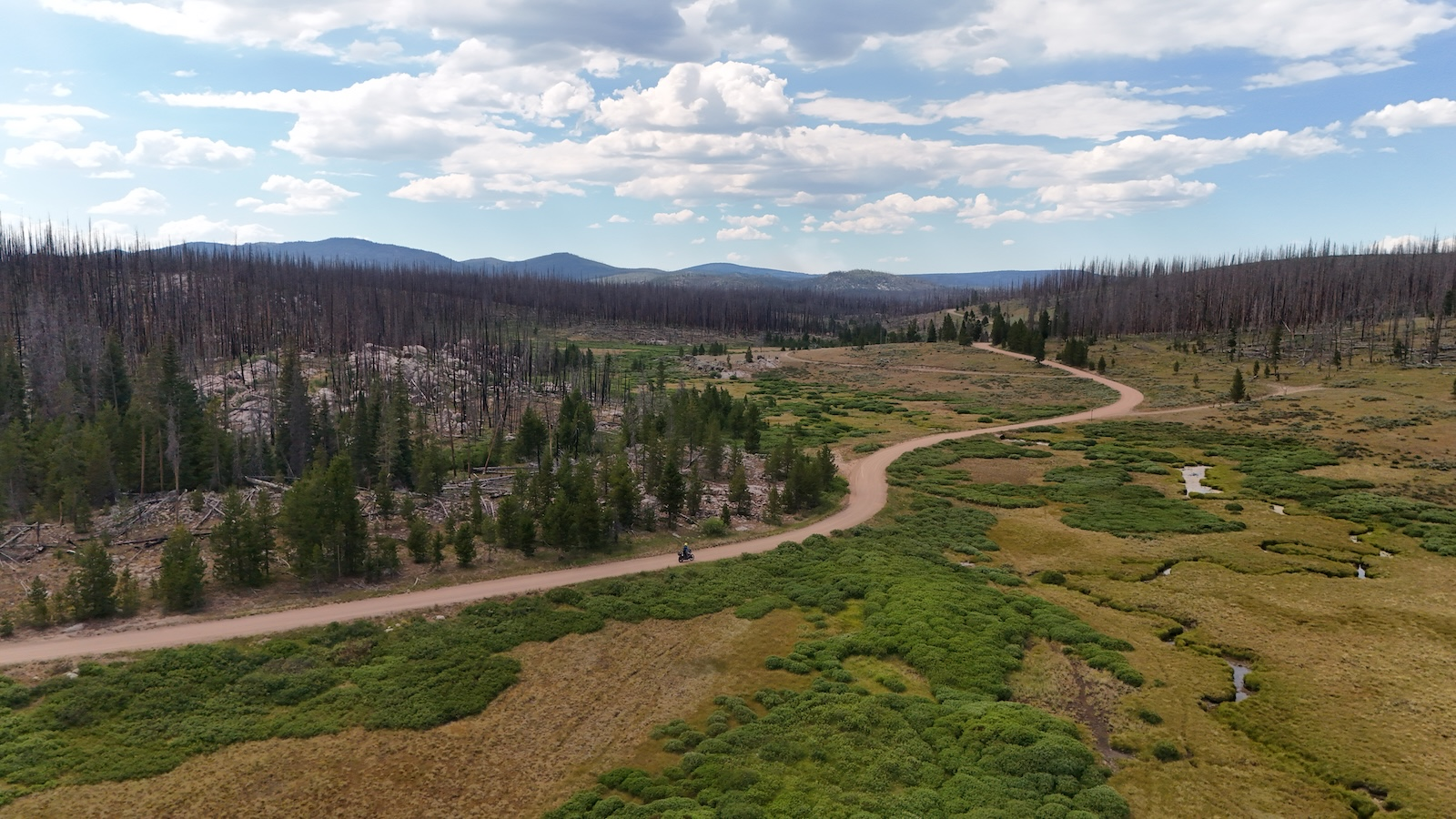 Aerial view of a dirt road winding through a green Colorado valley with burnt forest and mountains beyond