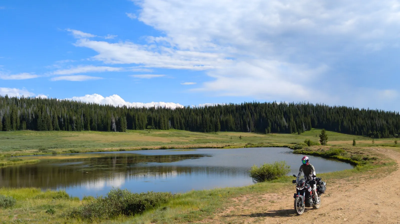 NastyNils in light adventure riding gear standing next to the loaded Aprilia Tuareg 660 at a Colorado mountain lake