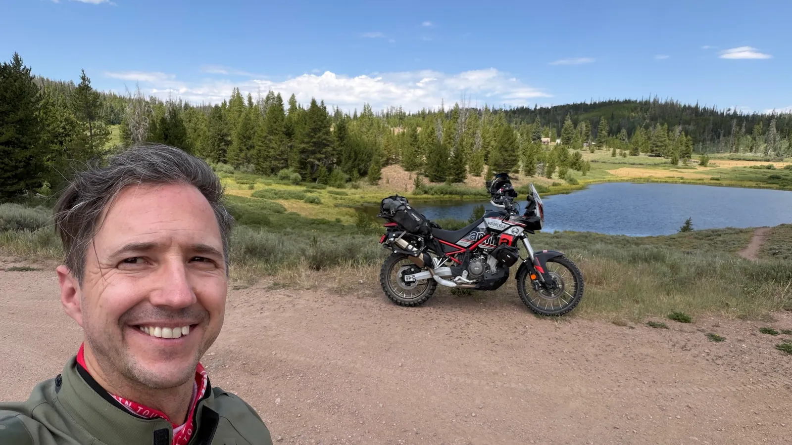 NastyNils smiling in the foreground with the loaded Aprilia Tuareg 660 parked at a Colorado alpine pond behind him