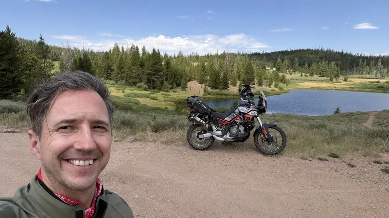 NastyNils in riding gear with the loaded Aprilia Tuareg 660 behind him at a Colorado mountain lake