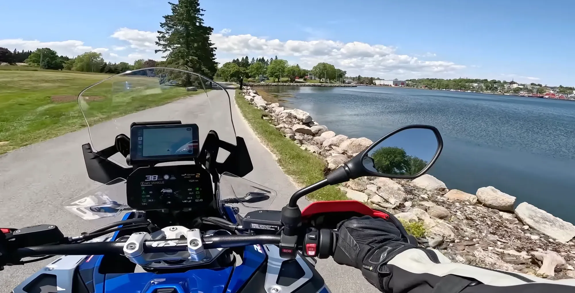 First-person onboard view from a BMW R 1300 GS Adventure, the round TFT dash glowing "My Vehicle" as the bike tracks a quiet waterfront road past a stone breakwater and a harbour town across the inlet.