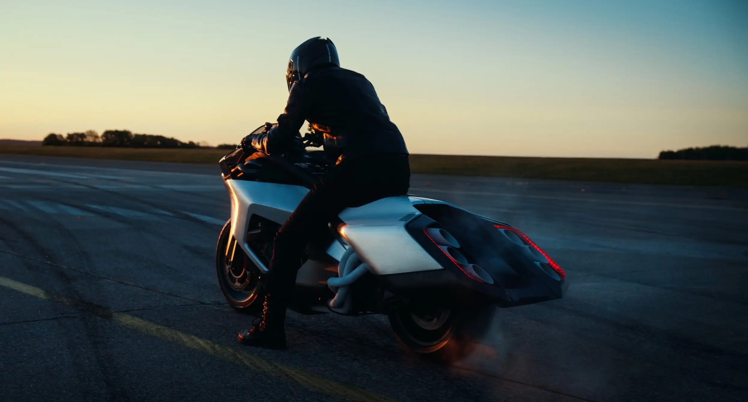 BMW K 18 Vision concept motorcycle on tarmac at sunset, rider in black gear seated low, exposed single-sided swingarm and minimalist sculpted bodywork visible, side-on shot at golden hour.