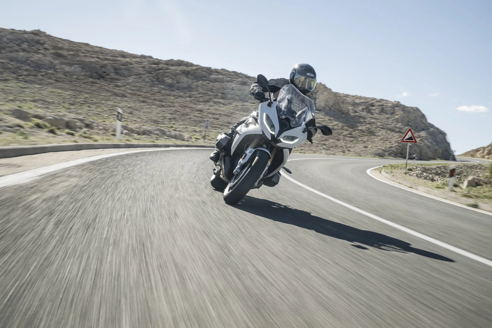 NastyNils riding a BMW R 1250 RT through a winding mountain road in Croatia, leaning hard through a right-hand curve. Rocky terrain and sparse vegetation visible in the background. Full leathers, full-face helmet, daylight conditions with clear sky. Road surface is dry asphalt, motion evident from lean angle and road positioning.