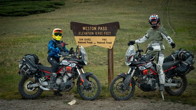 Two riders in full adventure gear beside their loaded Aprilia Tuareg 660s at the Weston Pass elevation sign