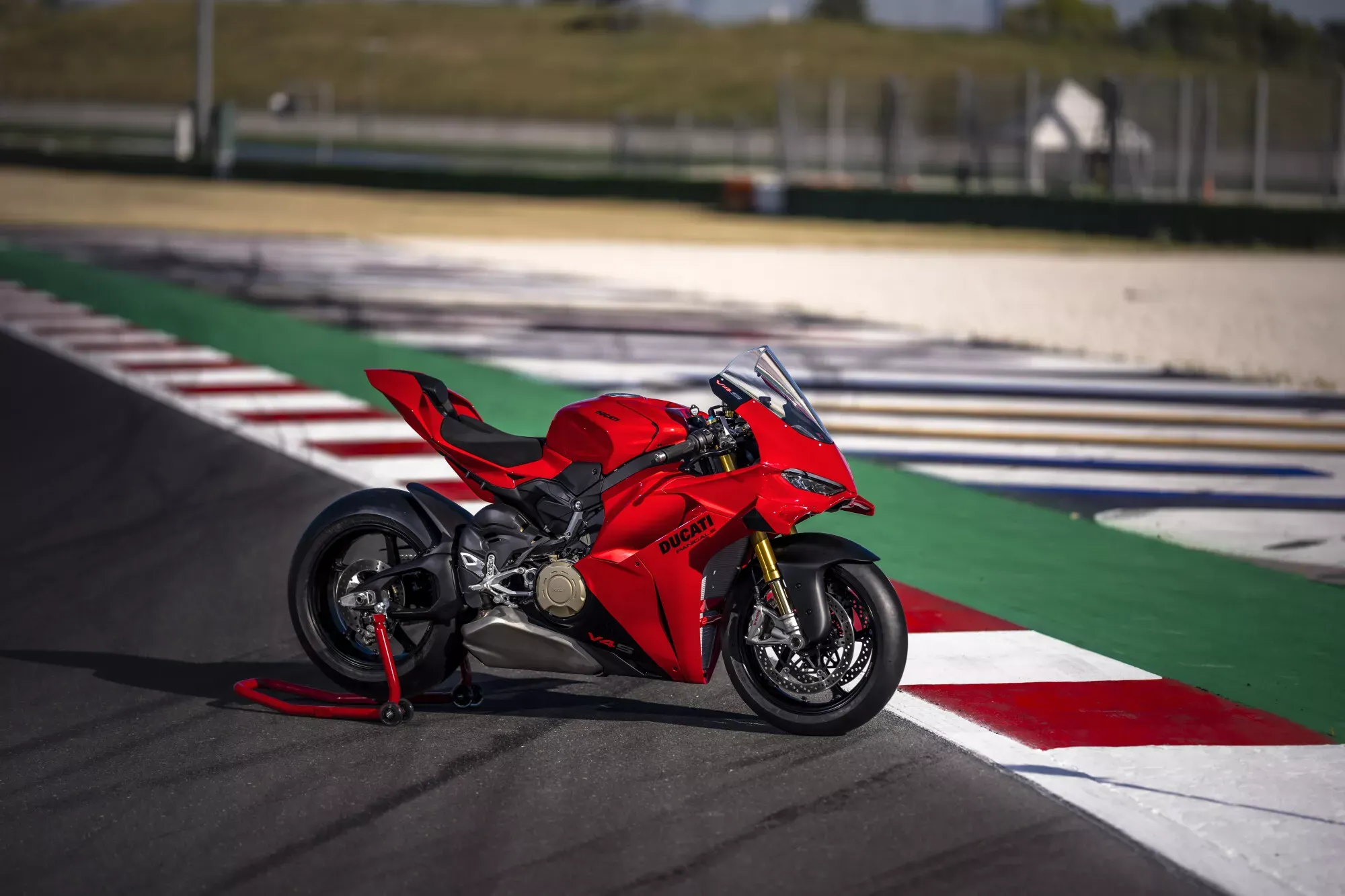 Red 2027 Ducati Panigale V4 standing on a red rear paddock stand at the edge of a race track, right side in a slight three-quarter front view, with red-and-white curbing, a green runoff and grandstands blurred behind it. No rider.