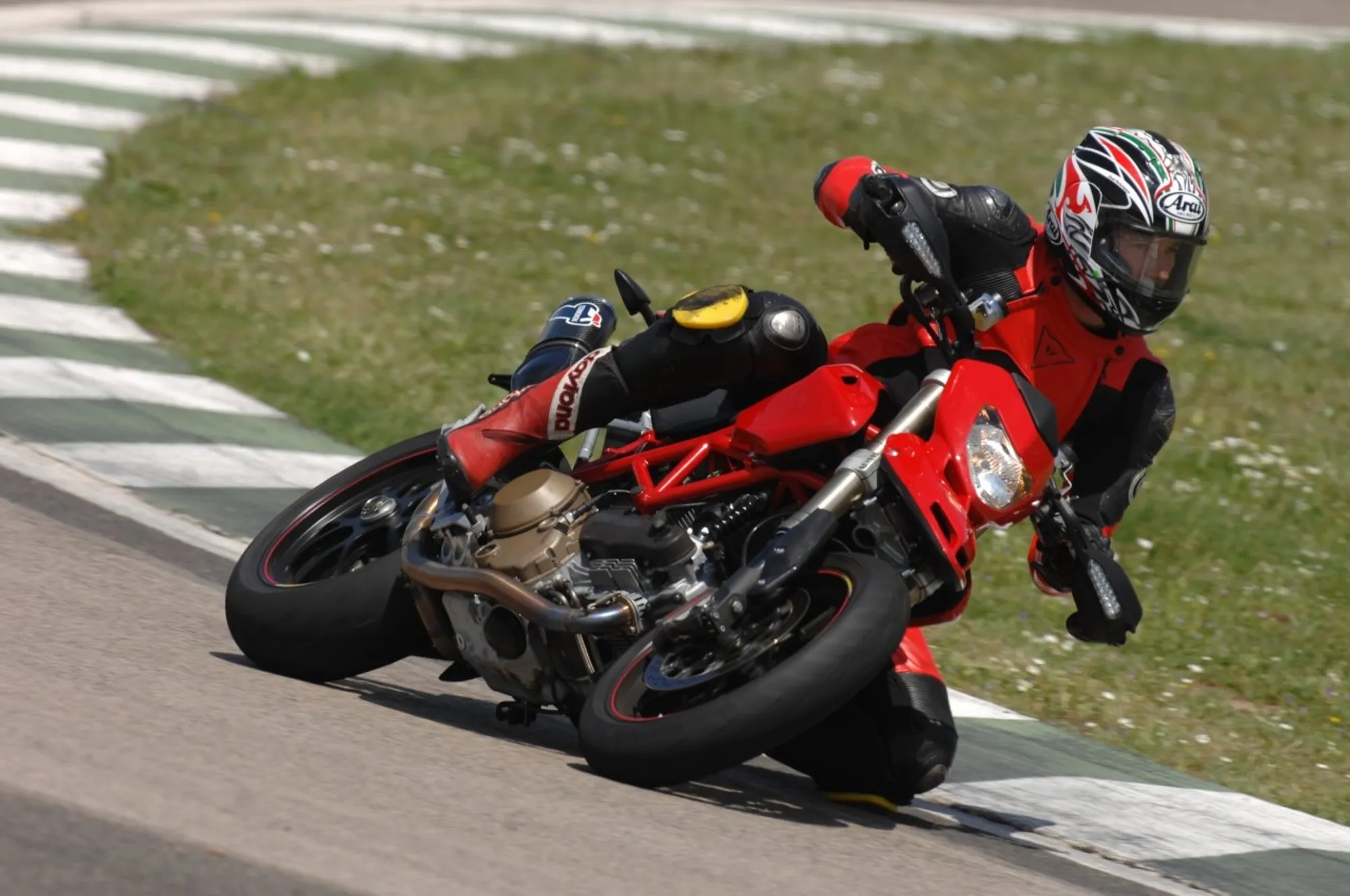 NastyNils riding a Ducati Hypermotard on a racetrack, leaning hard through a corner. The bike is red with black accents, positioned mid-corner at significant lean angle. He wears red and black leathers with a full-face helmet. Track setting visible with grass infield and white road markings. Daylight, clear dry conditions, high-speed cornering maneuver.