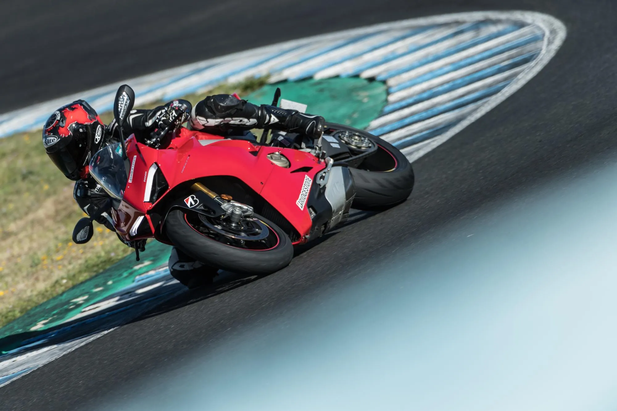 NastyNils cornering hard on a red Ducati Panigale at a racetrack, knee down through a left-hand bend. Heavy lean angle visible, with the rider in full leathers and a red full-face helmet. Bridgestone branding clearly visible on the bike's fairings. Dry asphalt, painted curbs and run-off area in the background. Daylight, sharp shadows.