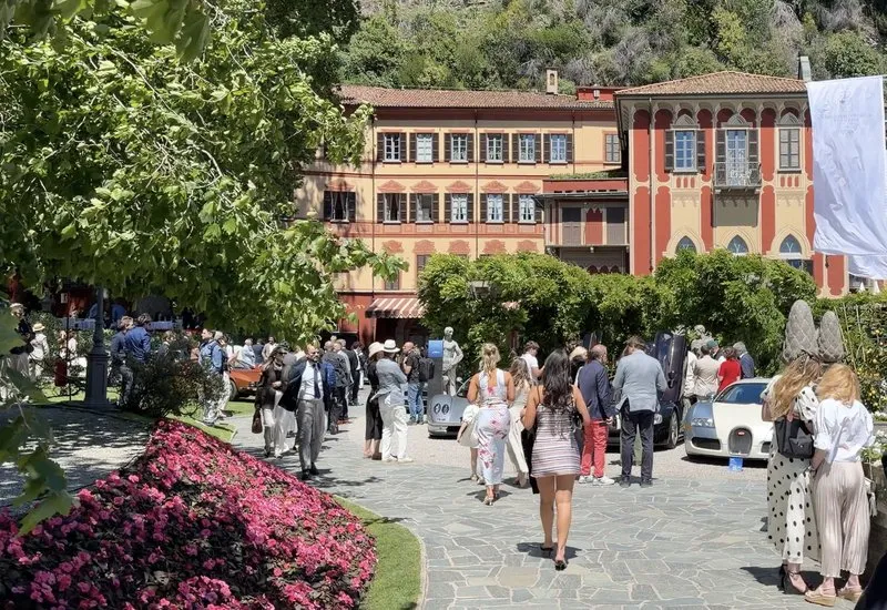 Elegant garden gathering at the Concorso d’Eleganza Villa d’Este on Lake Como, well-dressed attendees mingling between flowerbeds and classical statues with concours-class vehicles displayed under shaded loggia.