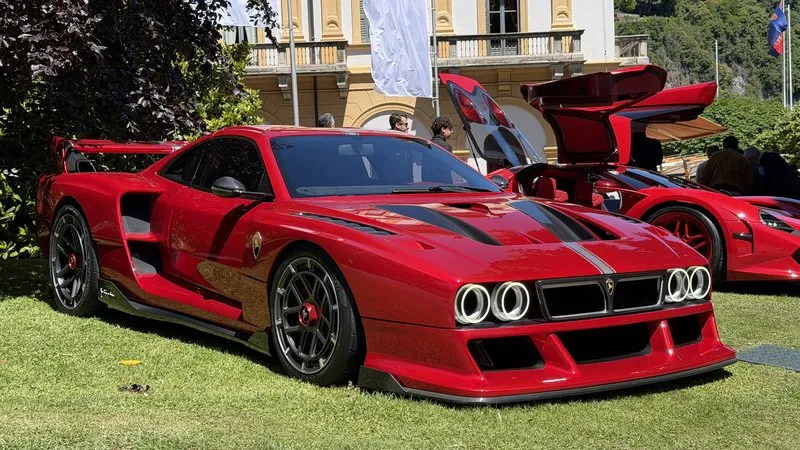 Red mid-engine supercar with twin round headlight pairs displayed on manicured grass in front of a yellow villa at Concorso d’Eleganza Villa d’Este on Lake Como, gullwing doors and red rim accents visible.